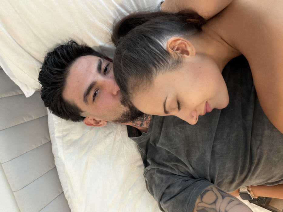 An overhead photograph captures two individuals, an adult male and an adult female, in a close embrace while lying in a bed. This scene is situated within a bedroom in Bangkok, Thailand.
The male, positioned behind the female, has dark hair styled with a fade, a beard, and visible tattoos on his neck and left arm. He is wearing a dark grey, subtly textured t-shirt. His eyes are open, directed towards the viewer. His left arm, featuring additional tattoos, rests on the bed.
The female is situated in front of and embraced by the male, resting her head against his upper chest or shoulder. Her dark hair is pulled back into a ponytail. A small, metallic stud piercing is visible on her right nostril. Her eyes are closed, suggesting repose or sleep. Her visible left arm wears a light-colored watch strap and a beaded bracelet. Her right arm is wrapped around the male. She appears to be shirtless or wearing minimal upper body clothing, with her bare shoulder prominent.
Both individuals are on white bedding, which includes a white pillow beneath the male's head. A light grey, vertically padded headboard is visible in the upper background of the frame. The ambient lighting is bright and even, consistent with an indoor setting.