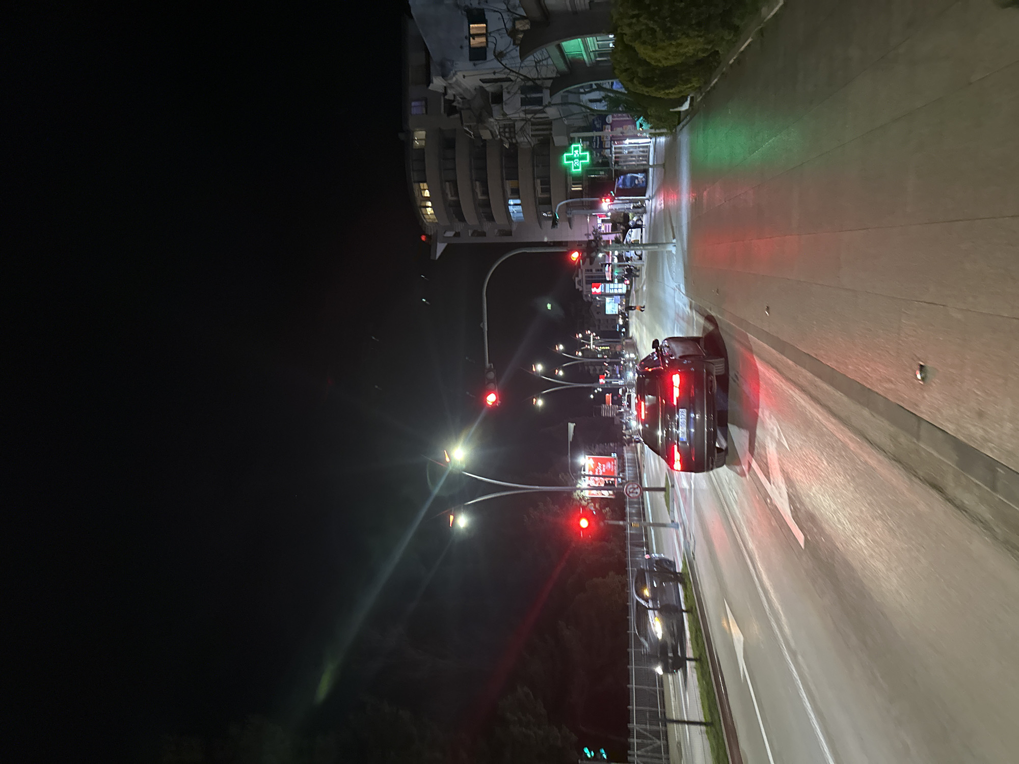 The image shows a nighttime street scene in Budva, Montenegro. The view is oriented along a roadway featuring two lanes, with a car traveling in the direction of the viewer. Red traffic lights are visible overhead. A green cross is seen above what appears to be a pharmacy. Buildings line the road on one side, with dark vegetation visible on the other. Car taillights and streetlights illuminate the scene, creating reflections on the wet pavement. A number plate is also visible.