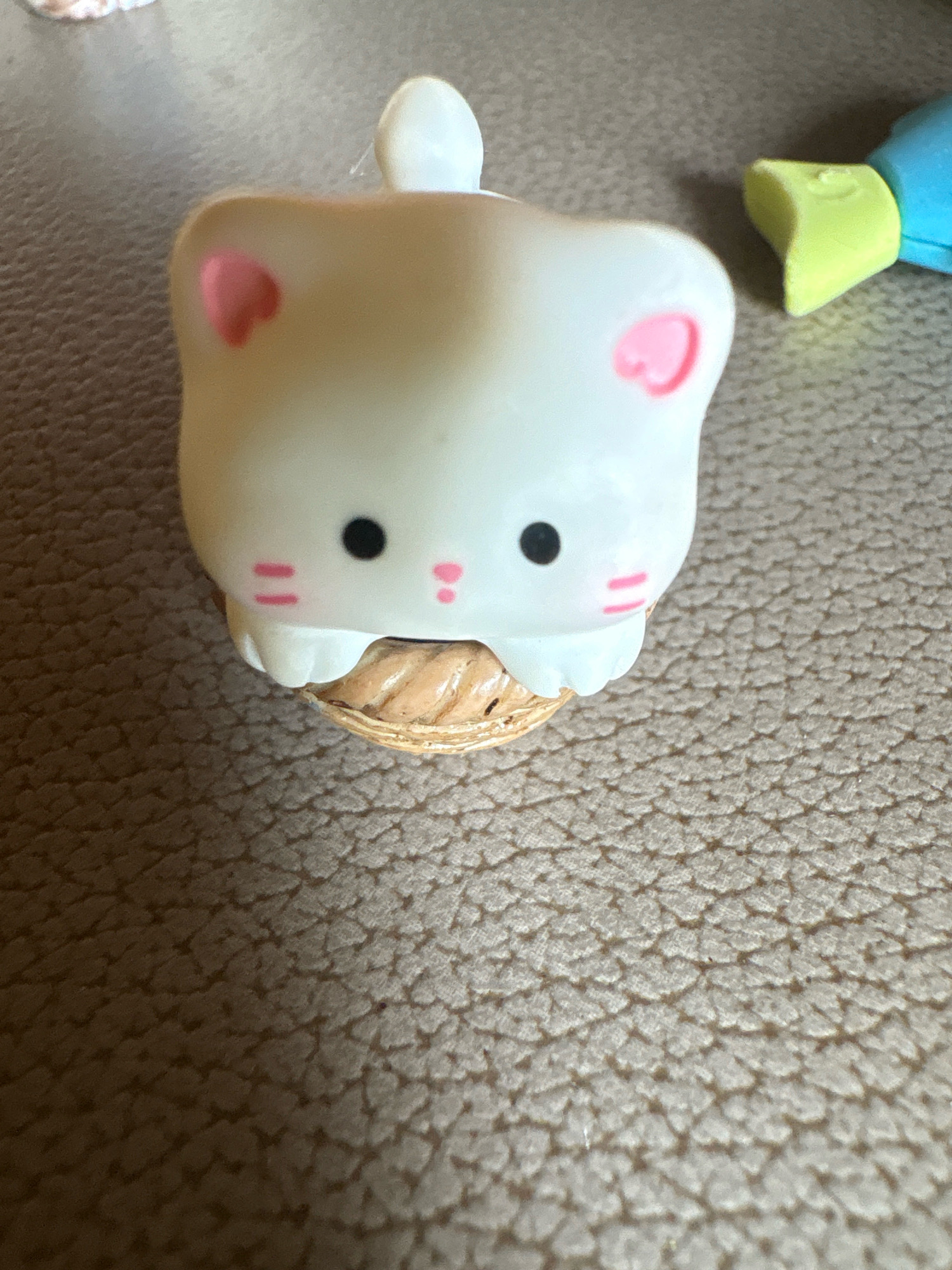 A close-up, high-angle shot shows a white cat figurine with pink heart-shaped ears and blush marks on its cheeks. It has black dot eyes and pink line markings below its eyes, resembling a mouth. The figurine sits atop a miniature woven basket. To the right of the figurine, a yellow and blue object, possibly a toy or part of another figurine, is visible. The background consists of a textured, brown surface. The scene is not set in Vagator, India.