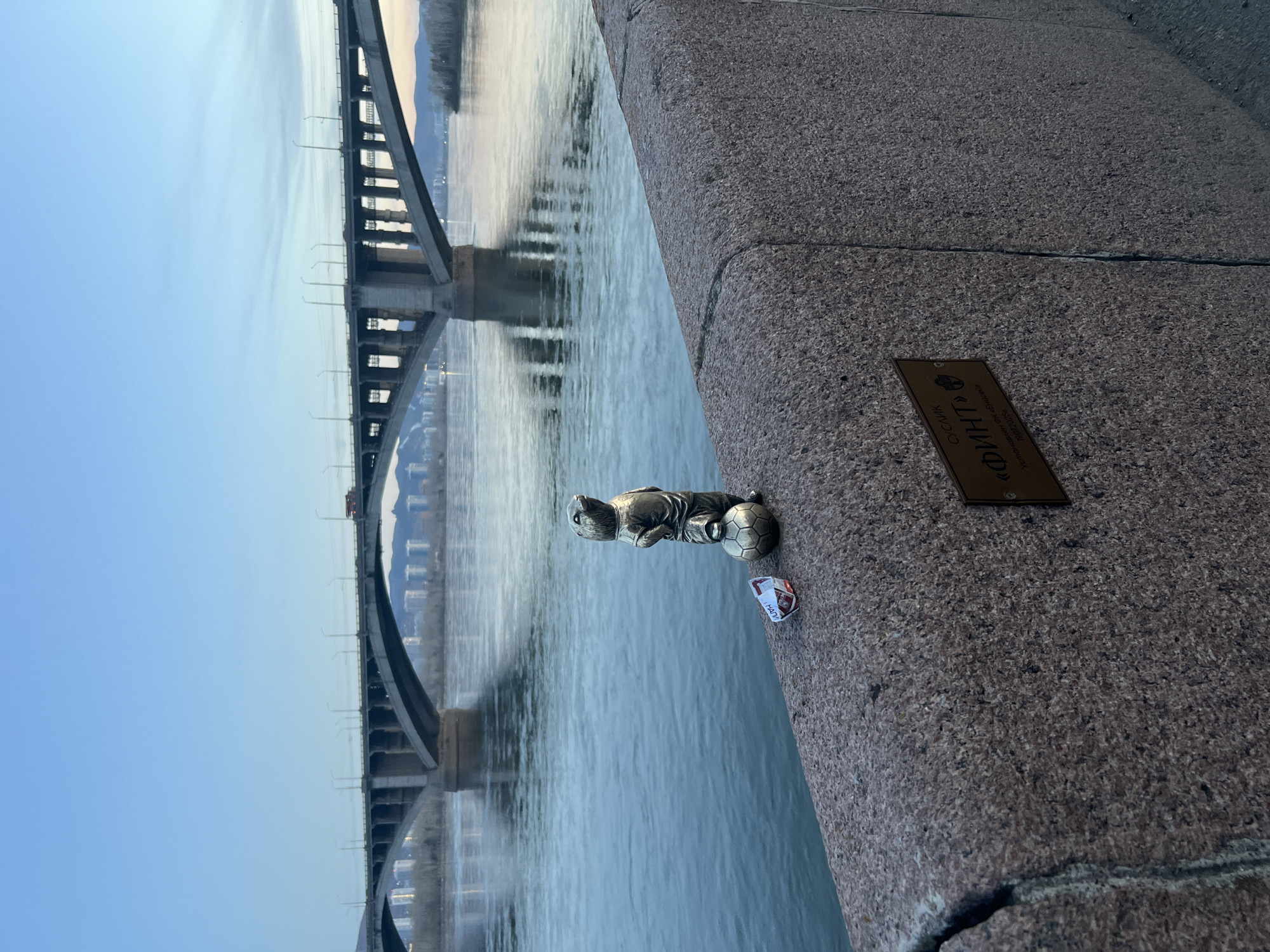The image captures a tranquil scene overlooking a wide river, likely the Yenisei in Krasnoyarsk, Russia, as suggested by the visible text and provided context. In the foreground, a sturdy granite or concrete embankment forms a diagonal line, separating the viewer from the water.
On this embankment, a small metallic figurine is prominently displayed. It depicts a stylized animal, possibly a bear or similar creature, wearing a sports jersey and shorts, standing next to a miniature soccer ball. Near the figurine, a crumpled piece of white and red paper, possibly a candy wrapper, lies on the stone surface.
Adjacent to the figurine, a small rectangular plaque, appearing to be made of brass or bronze, is affixed to the embankment. The text on the plaque is in Cyrillic, reading: "ОКСЮИК" (OXYUIK), followed by "ФИНТ" (FINT), which means "feint" or "trick," a term commonly used in sports. Below that, it states "установлен от Енисея" (ustanovlen ot Yeniseya), translating to "installed by Yenisei" or "from Yenisei," confirming the location by the Yenisei River.
Beyond the embankment, the calm, grey-blue water of the river reflects the soft light of the sky and the structures along its banks. A large, multi-arched bridge, possibly a railway or vehicle bridge, spans the river in the midground, its dark silhouette mirrored in the water below. Its structure suggests a robust construction of concrete and steel.
In the distance, low-lying hills or mountains are visible on the horizon. The sky is clear, displaying a gentle gradient from a pale blue at the horizon to a deeper blue overhead, indicative of either early morning or late afternoon lighting. The overall mood is peaceful and reflective.