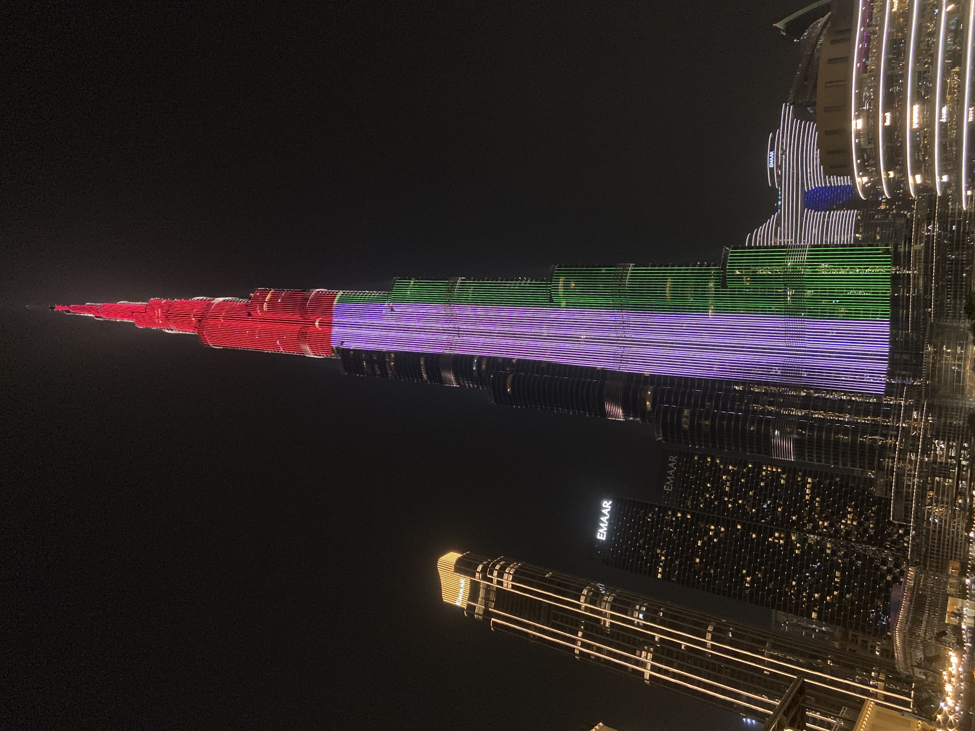 The image shows the Burj Khalifa in Dubai illuminated with the colors of the United Arab Emirates flag: red at the top, followed by green, purple, and black. The night sky is dark, contrasting with the bright lights of the buildings. Other skyscrapers are visible, including one with the word "EMAAR" on its facade. The buildings are lit with white and gold lights.