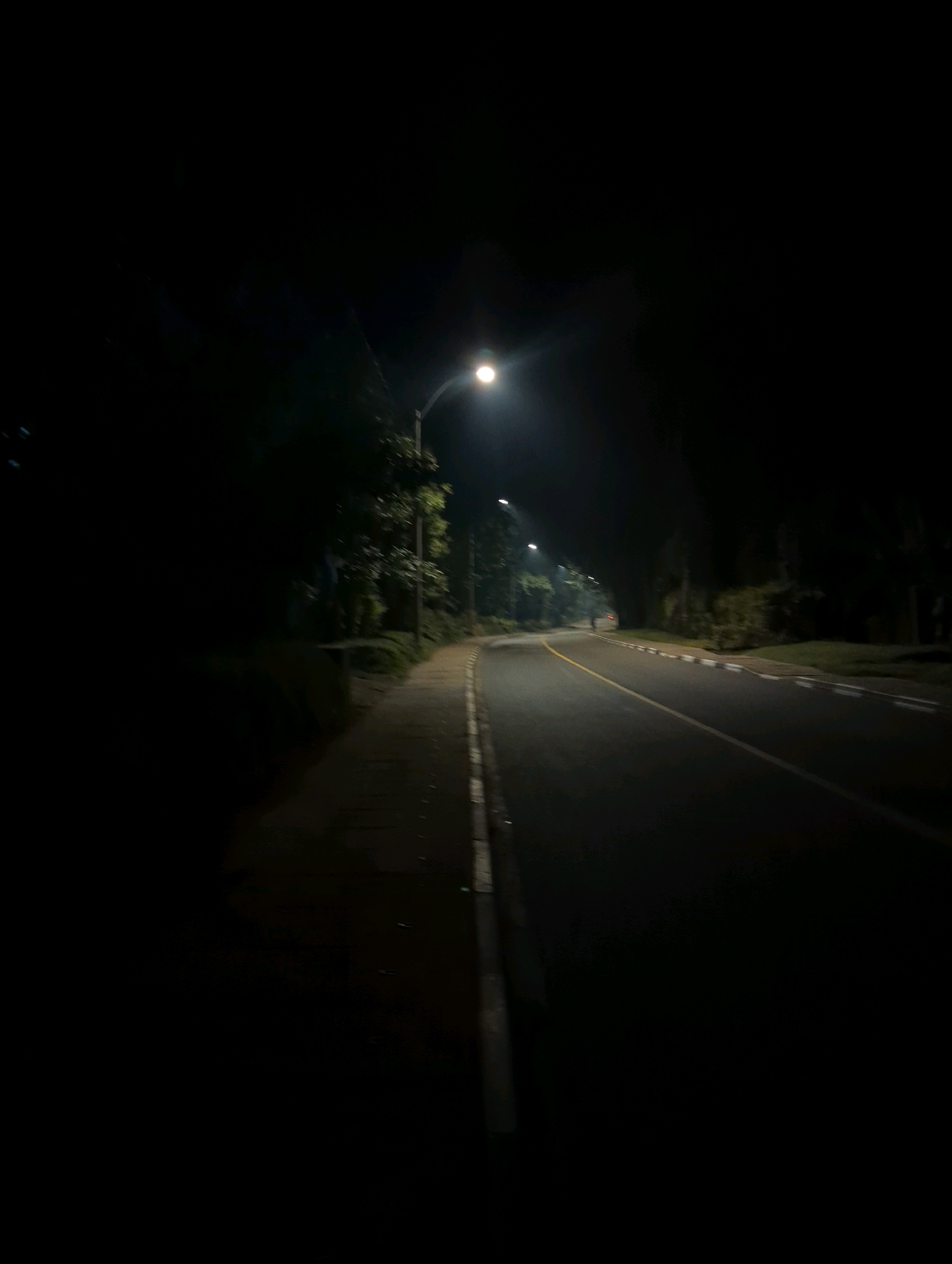 The image depicts a paved road at night in Musanze District, Rwanda. The road is illuminated by streetlights, casting a dim glow on the asphalt. A sidewalk runs alongside the road, separated by a curb. Trees and vegetation line the sides of the road, their forms obscured by the darkness. The scene is empty of people and vehicles, creating a sense of solitude. The overall atmosphere is quiet and serene.