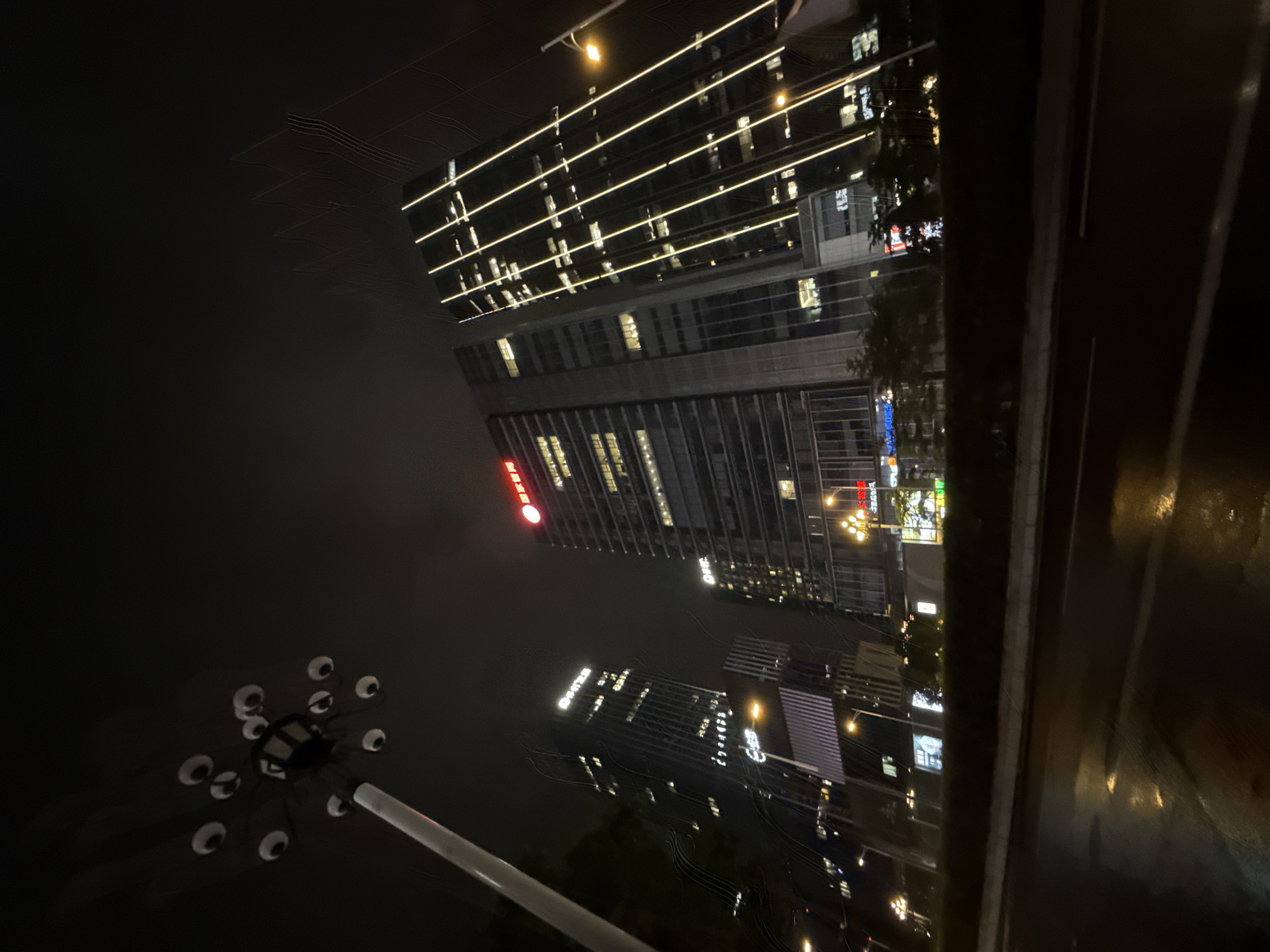 The image captures a nighttime view of modern skyscrapers in Tianhe District, China. A streetlamp with multiple lights resembling eyes is visible in the foreground on the left, casting an eerie glow against the dark sky. Several tall buildings dominate the background, their facades illuminated by rows of vertical yellow lights and numerous lit windows, indicating activity within. One building displays red Chinese characters at its top. The street in front of the buildings appears wet, reflecting the lights, suggesting recent rain. The overall scene conveys an urban, metropolitan atmosphere at night.