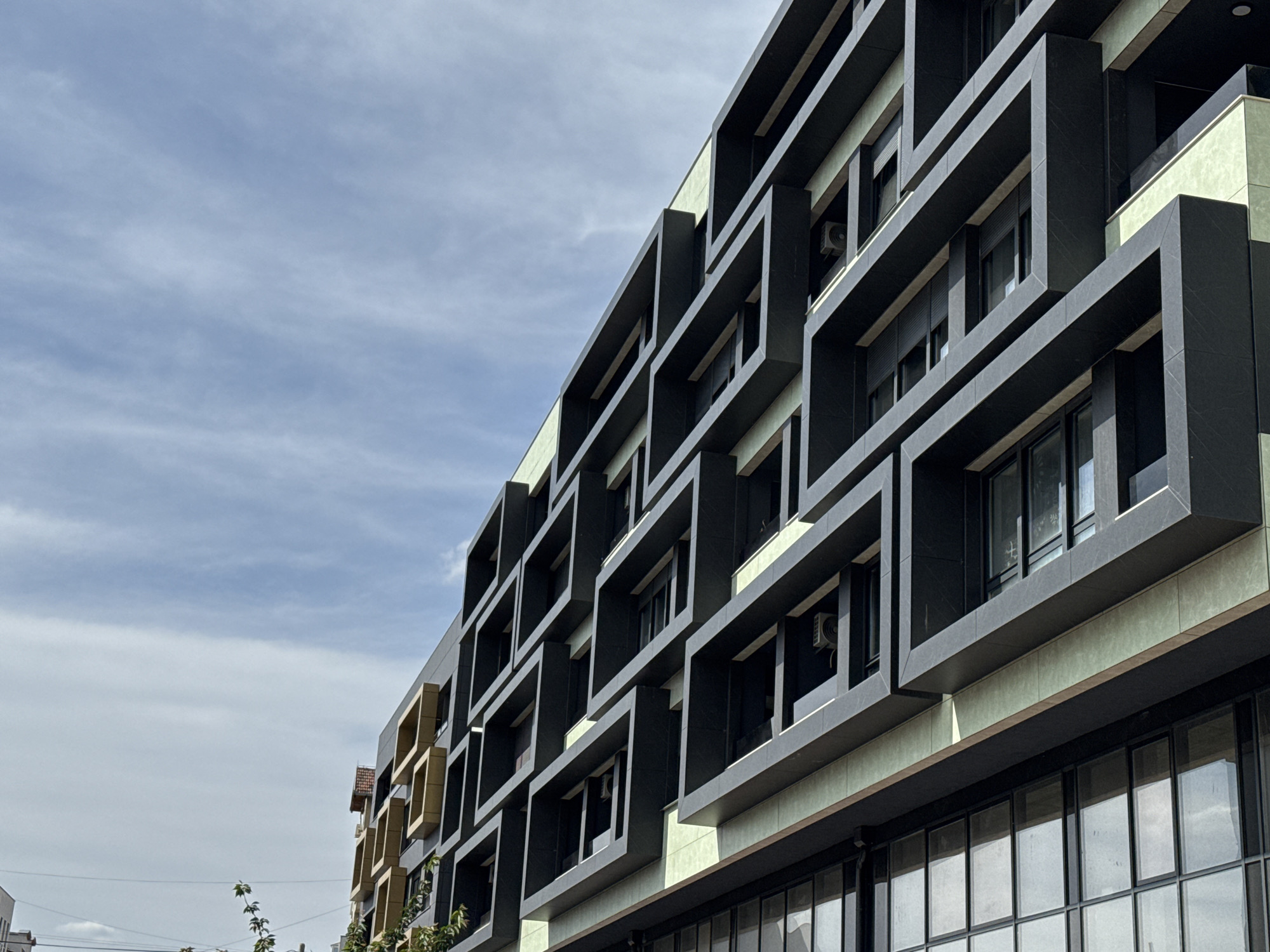 A low-angle medium shot reveals the facade of a modern building in Novi Sad, Serbia, characterized by an orthogonal grid of protruding, dark-colored rectangular frames surrounding windows. The building, potentially a residential or commercial structure, displays a pale green surface between the dark frames. Large windows cover the ground floor. Above, each level features windows set within the protruding frames. The sky is partly cloudy with a light blue hue. Other buildings are faintly visible in the periphery of the shot.