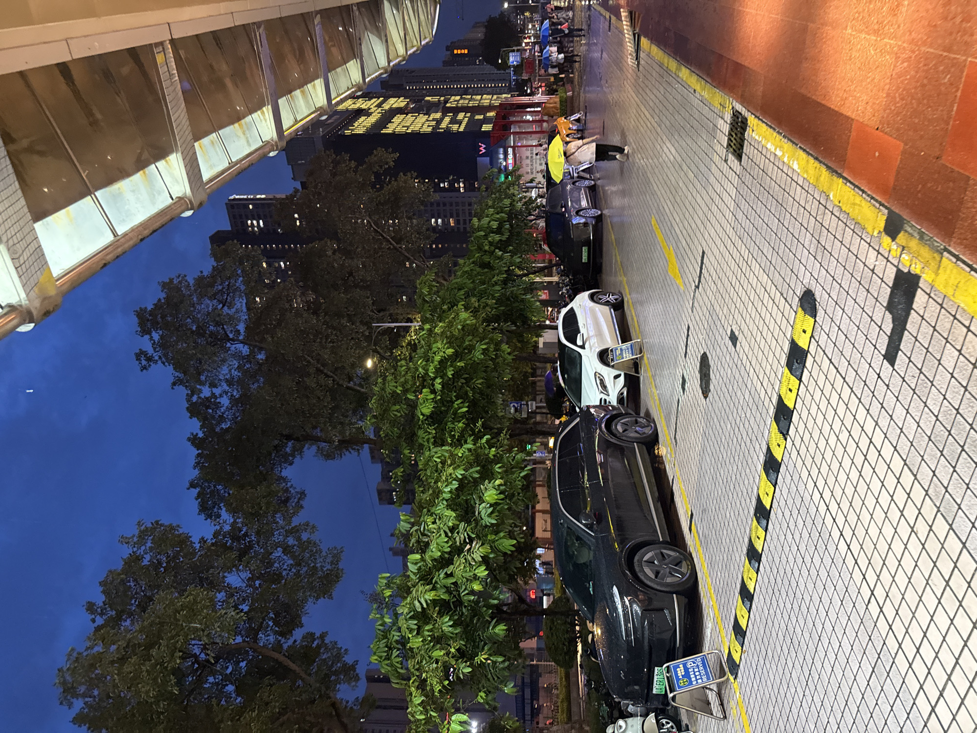 This media file captures a street scene at dusk in Tianhe District, China. The perspective is from a covered walkway looking down a tiled street lined with trees and buildings. Several cars are parked along the street, including a dark-colored sedan and a white car in the foreground. A few people are visible, some holding umbrellas, suggesting it might be raining or has recently rained, as the tiles appear wet. One person is wearing a bright yellow jacket, and another a blue umbrella. The background features a tall building with illuminated windows and a large sign with yellow text. The overall mood is urban and somewhat subdued, typical of early evening in a city. Visible text includes Chinese characters on the building sign and license plates on the cars, one of which reads "粤EPR365". The time of day is evident from the twilight sky, a deep blue indicating the transition from day to night.