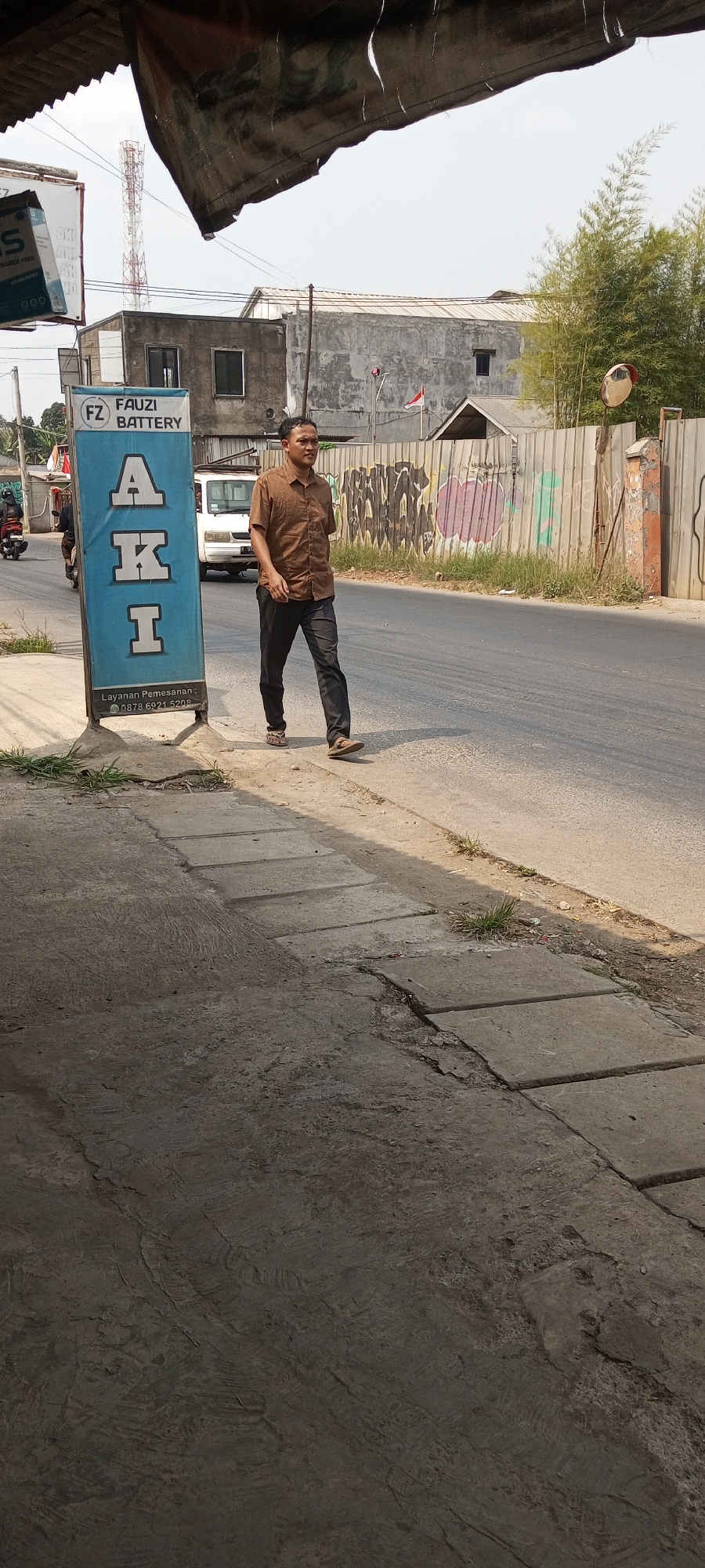 A man in a brown collared shirt and dark pants walks on a sidewalk next to a road in Setu, Indonesia. A blue sign with the words "FAUZI BATTERY AKI" is positioned on the sidewalk to his left. Behind the man, a white van is parked on the side of the road, and a metal fence with graffiti runs along the roadside. A tall tower is visible in the distance, and lush green trees are present on the right side of the image. The sky is clear and bright, indicating a sunny day. The sidewalk is paved with concrete slabs and shows signs of wear and tear.