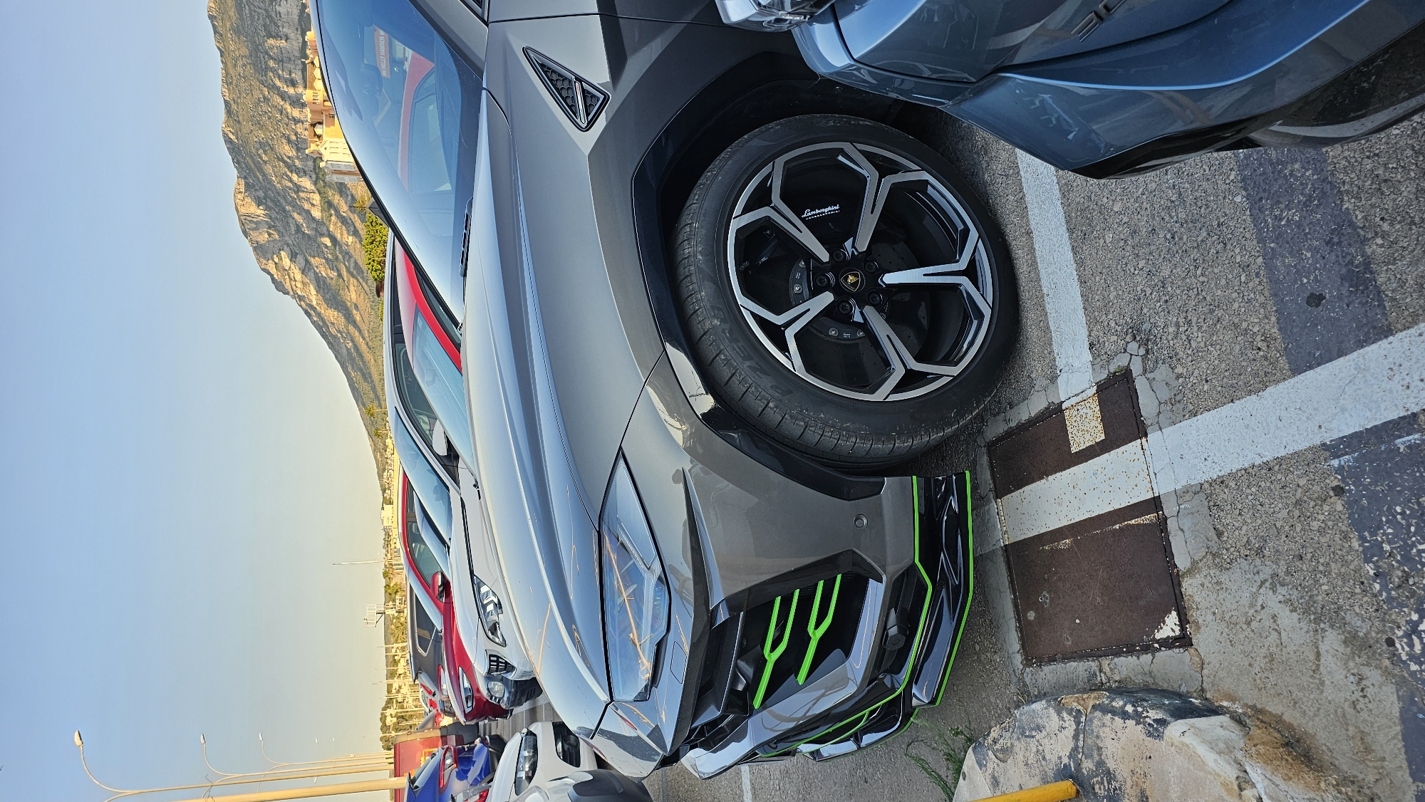 The image depicts a row of parked cars in an outdoor lot with a mountainous backdrop. The foreground is dominated by the front quarter of a dark grey Lamborghini Urus, distinguished by its aggressive styling, green accents on the front bumper and side skirts, and distinctive multi-spoke wheels. Behind it, other vehicles are visible, including a red car and a white car. The parking lot surface is asphalt with painted white lines. In the background, buildings and a rugged mountain range under a clear blue sky suggest a Mediterranean setting. The lighting indicates it is late afternoon or early evening. The scene is devoid of any people. Based on the visual cues of the distinctive mountain and coastal vegetation, this location is likely Dénia, Spain, specifically the area near the Montgó mountain.