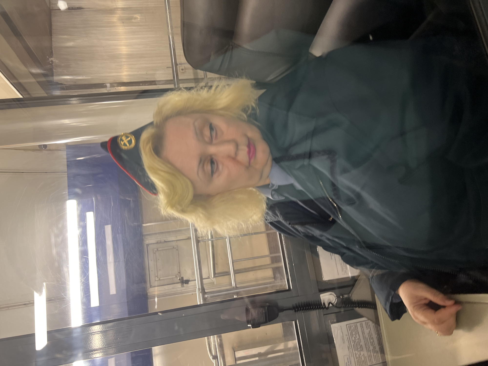 A woman in a dark green jacket and a dark cap with a gold emblem sits behind a glass window. The cap has a red band. The woman has blonde hair and pink lipstick. Behind the window are fluorescent lights, a metal structure, and paperwork.