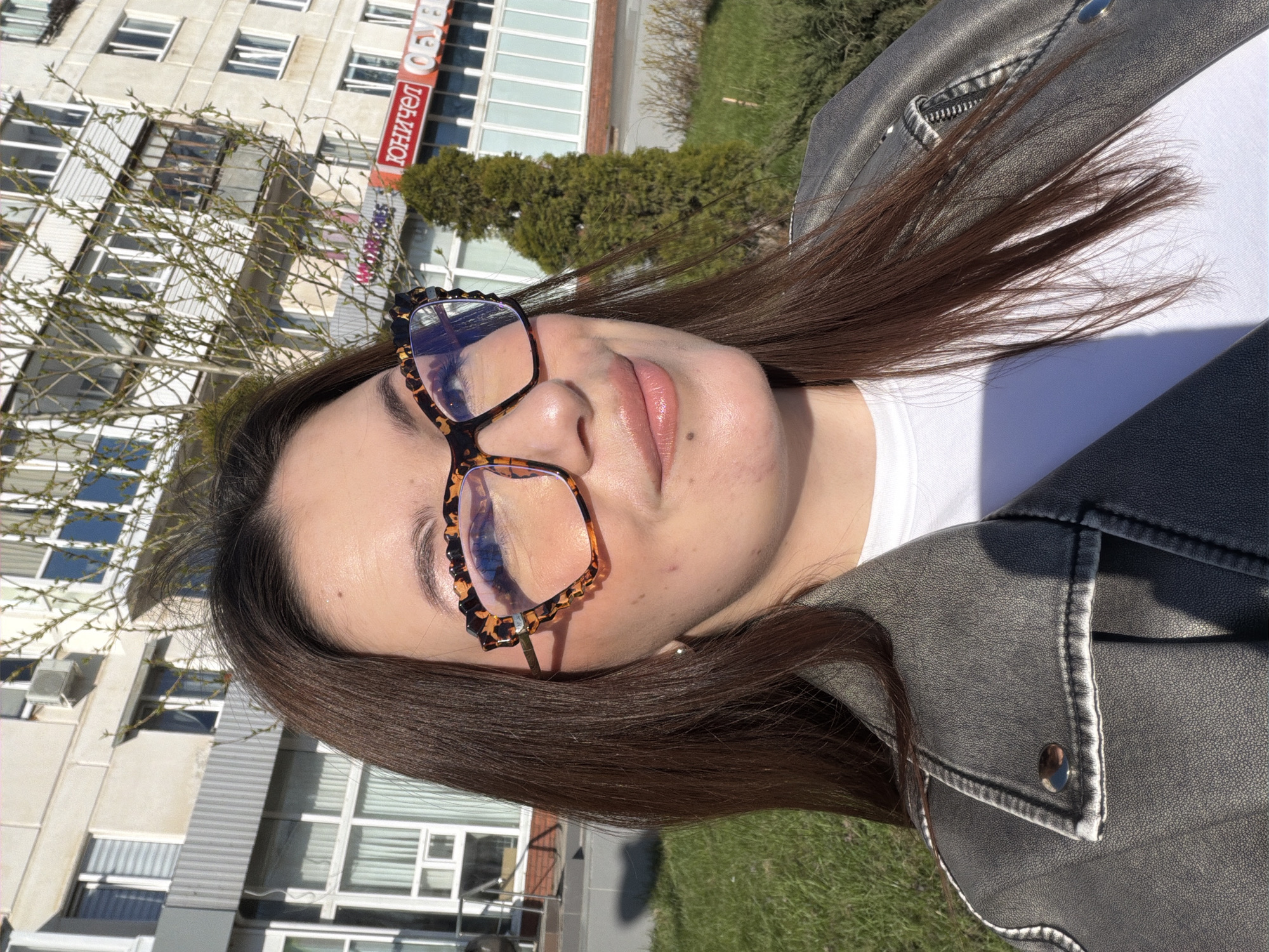 A woman is posing for a selfie outdoors in Chaikovsky, Russia. The woman has medium-length brown hair, fair skin, and is wearing tortoise-shell glasses with black detailing, a white T-shirt, and a gray leather jacket. The background shows a city setting, including a multi-story building, green grass, bushes, and the branch of a tree with buds. A sign with Cyrillic text (possibly "ЮНИЧЕЛ ОБУВЬ") is also visible on one of the buildings.