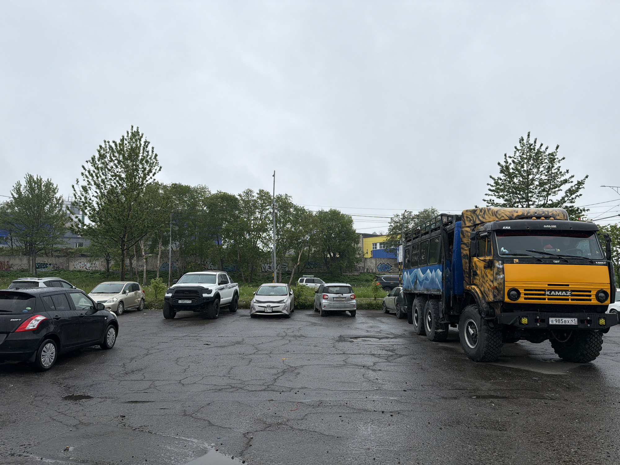 An outdoor scene in Petropavlovsk-Kamchatsky, Russia, depicts a parking lot on a cloudy day. Several vehicles are parked, including a black hatchback on the left, a white pickup truck, a silver compact car, and a larger truck with a yellow front and blue sides, decorated with a tiger pattern. The ground is wet and cracked, suggesting recent rain. In the background, there are trees, a building with graffiti, and telephone poles. The sky is overcast. No people are visible in the image.