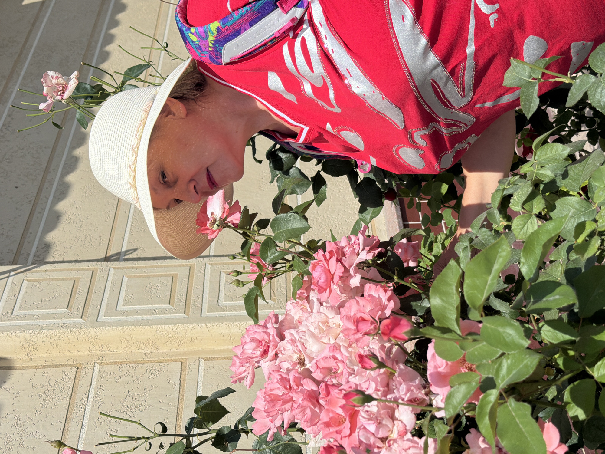 An older woman wearing a white straw hat and a bright red shirt with silver metallic accents is smelling a bush of pink roses. She is positioned in the right half of the frame, looking down and to her left, with her face partially obscured by the brim of her hat and shadows. The rose bush occupies the left and lower portions of the image, featuring numerous pink blooms and green leaves. The background consists of a textured, beige wall with square indentations. The scene is illuminated by bright sunlight, casting shadows from the foliage onto the wall and the woman's hat.