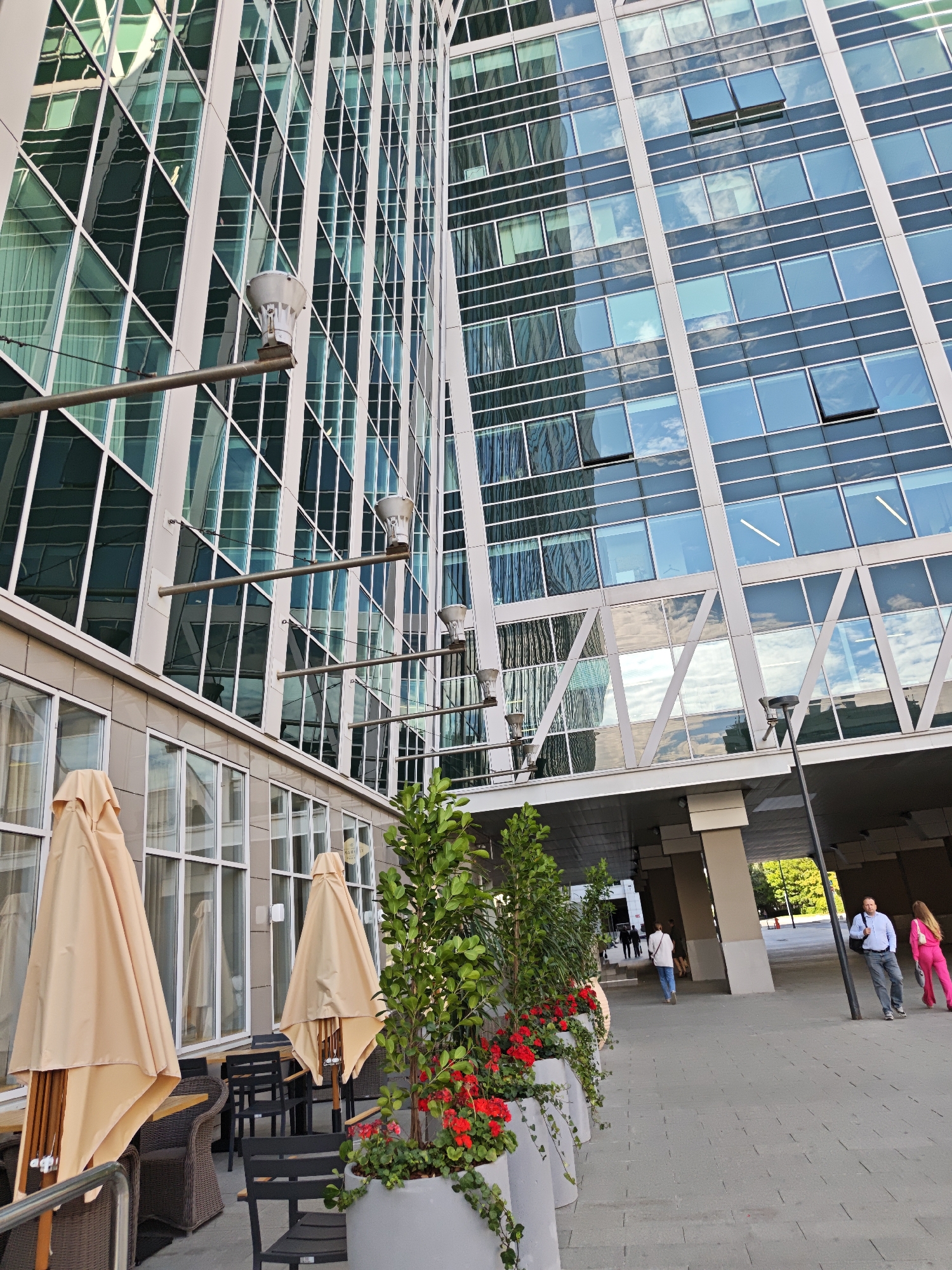 A street-level, eye-level shot captures the facade of a modern skyscraper in Moscow, Russia, with its reflective blue-tinted glass windows and white framing. The building's angular design is prominent, with sections appearing to twist and jut out. In the foreground, outdoor seating with tables and chairs is partially obscured by beige, folded umbrellas. Planters filled with lush green foliage and vibrant red flowers are arranged along the walkway. Several people are visible in the distance, walking under an elevated walkway that connects to the skyscraper. A man in a light blue shirt and dark pants, and a woman in a bright pink tracksuit are walking away from the camera. The environment appears to be an urban setting with trees and other buildings visible in the background.