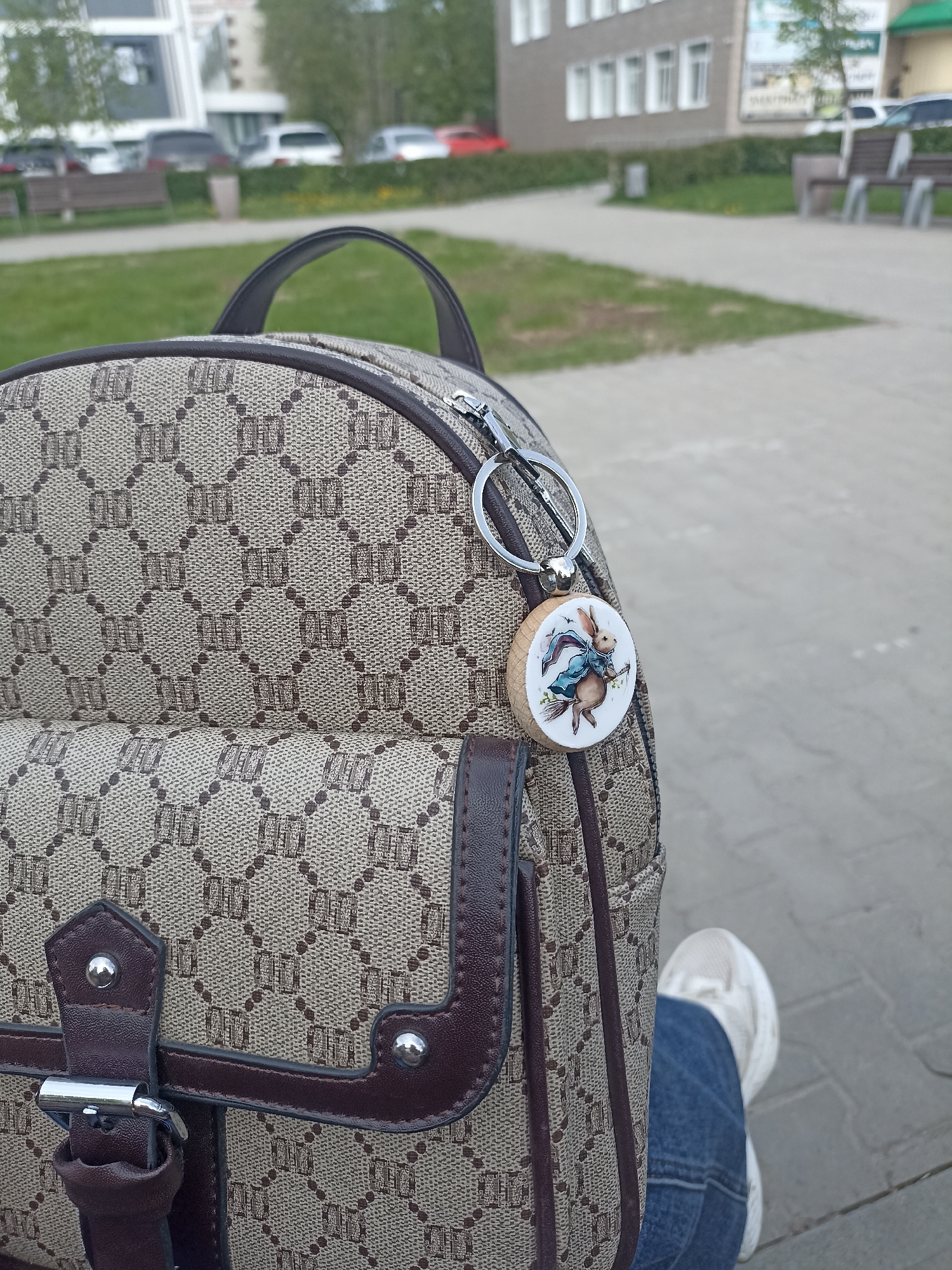 A close-up shot shows a person sitting outside on a paved surface, likely in a city park. The focus is on a brown patterned backpack with a small pocket and a round keychain featuring a whimsical illustration of a rabbit riding a broom. The person is wearing blue jeans and a white sneaker. In the blurred background, there are buildings, benches, trees, and cars, suggesting an urban environment.