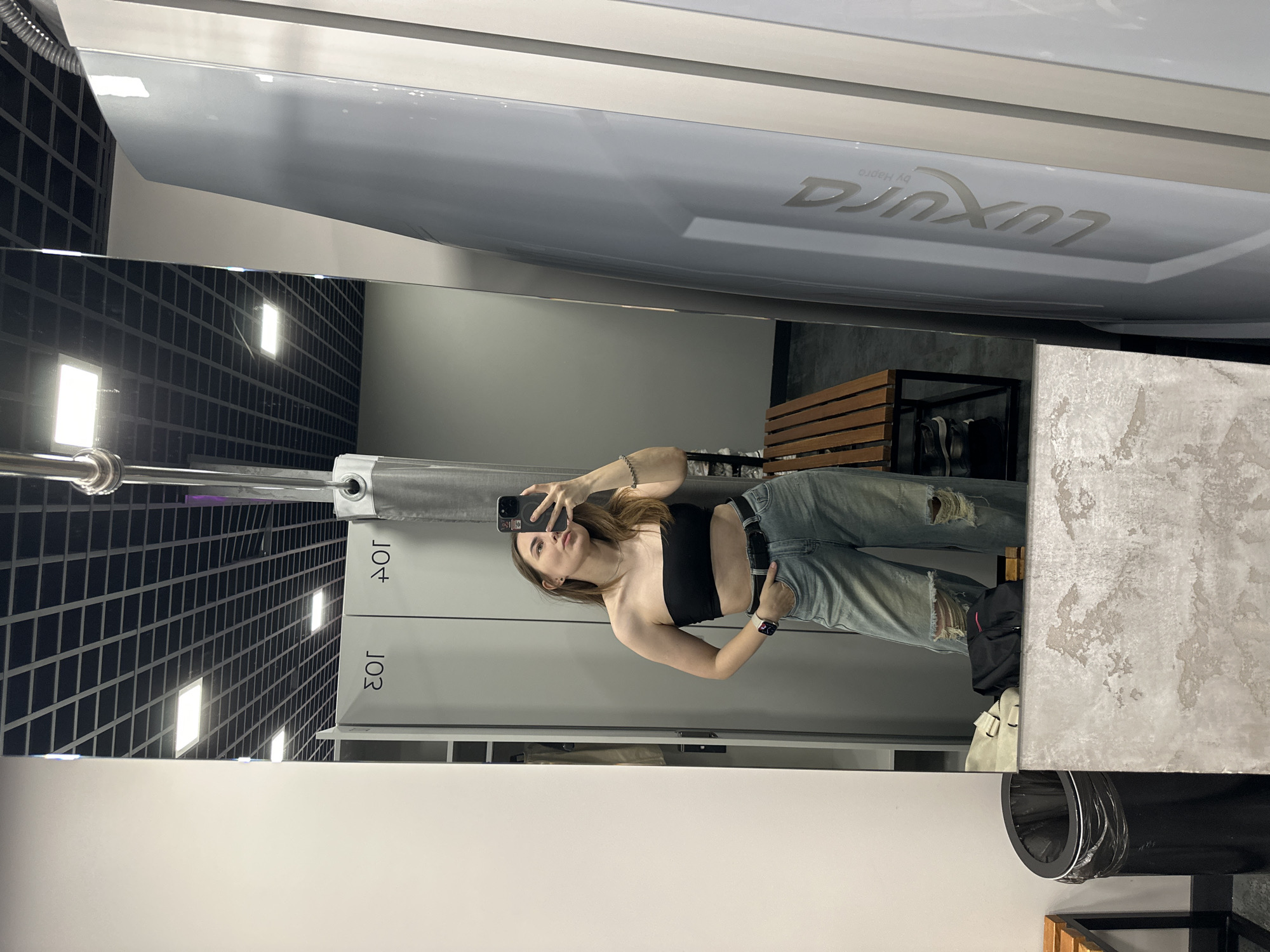 A young woman is taking a selfie in a mirror. She is wearing a black crop top and ripped jeans. A tanning bed with the "Luxura by Hapro" logo is visible above the reflection. The location appears to be a tanning salon, as indicated by the tanning bed and numbered lockers in the background. Based on the available information, it is not possible to definitively confirm that this photo was taken in Perm, Russia.