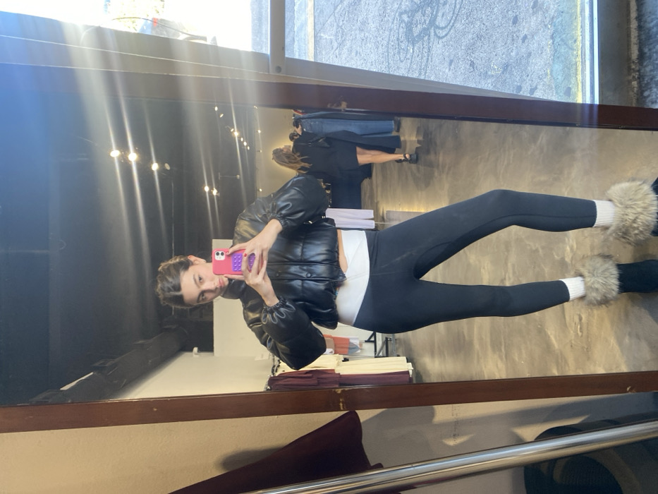 A young woman wearing a black puffer jacket, white top, and black leggings with furry ankle cuffs takes a mirror selfie with a pink phone. She is standing in what appears to be a clothing store, with racks of clothes visible in the background. Another woman is also visible in the background. The overall lighting is bright. The photo is taken at a slight angle.