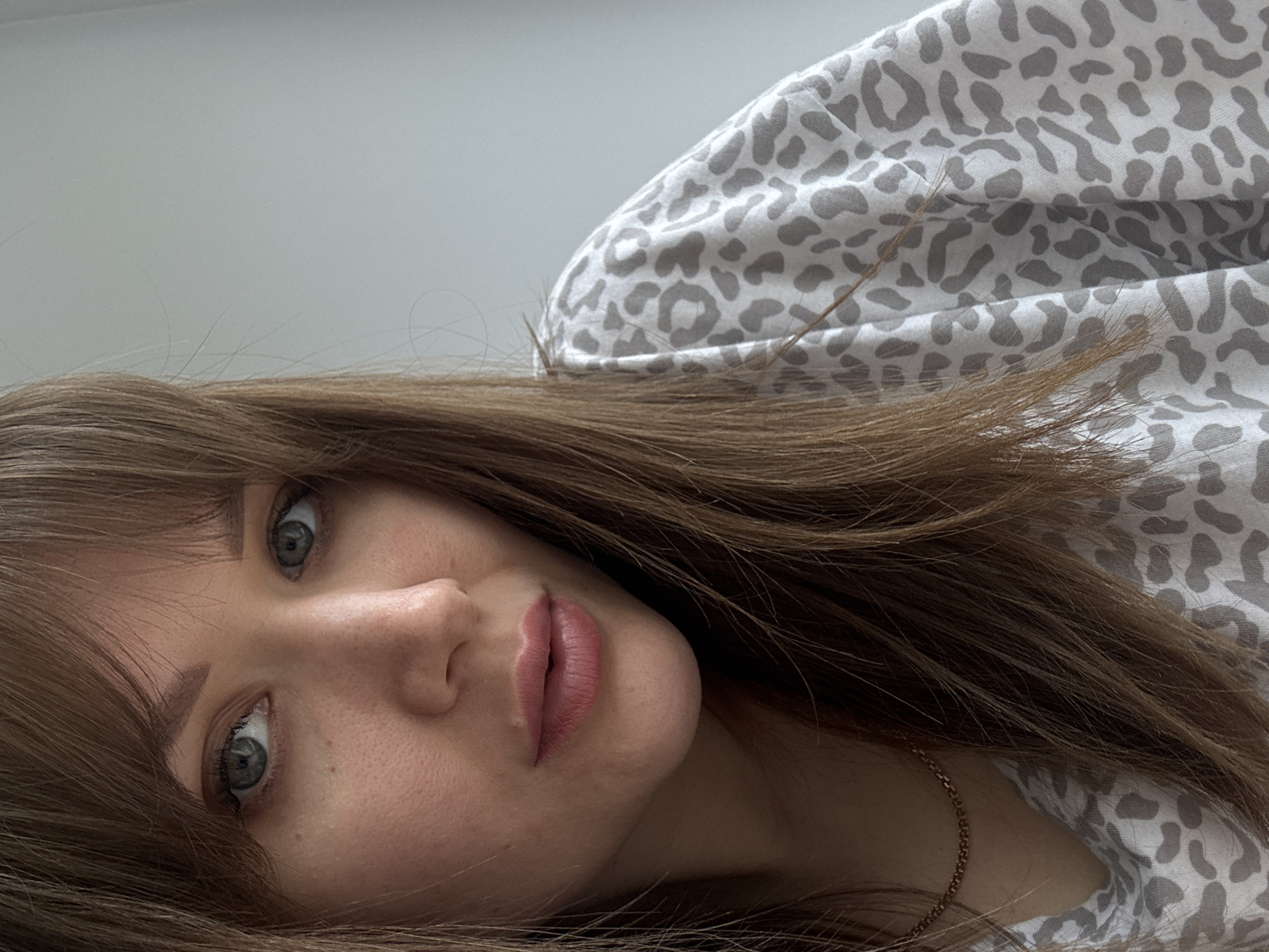 The media file presents a close-up, angled portrait of a young woman. Her head is tilted to the left, and the image is rotated counter-clockwise, giving the impression that she is lying down and looking towards the viewer.
**Subjects:** The primary subject is a young woman. She has long, straight, medium brown hair styled with bangs that fall across her forehead. Her eyes are a striking light blue-grey, wide open and directed towards the camera. Her facial features include a small nose and full, slightly parted lips, which appear to have a muted pink or nude lipstick applied. Visible freckles dot her nose and cheeks. A delicate gold chain necklace adorns her neck. She is wearing or partially covered by a garment featuring a light grey leopard print pattern on a white background.
**Scene/Setting:** The background behind her head is a plain, light grey or white surface, suggesting an indoor setting such as a room with a light-colored wall or ceiling.
**Activity or event taking place:** The woman is posing for a close-up portrait. No specific activity or event is depicted beyond this.
**Notable details:** The lighting is soft and even, consistent with indoor ambient light. Her expression appears neutral, calm, and contemplative. The time of day and external weather conditions are not discernible from the image.
**Visible text:** There is no visible text in the media file.