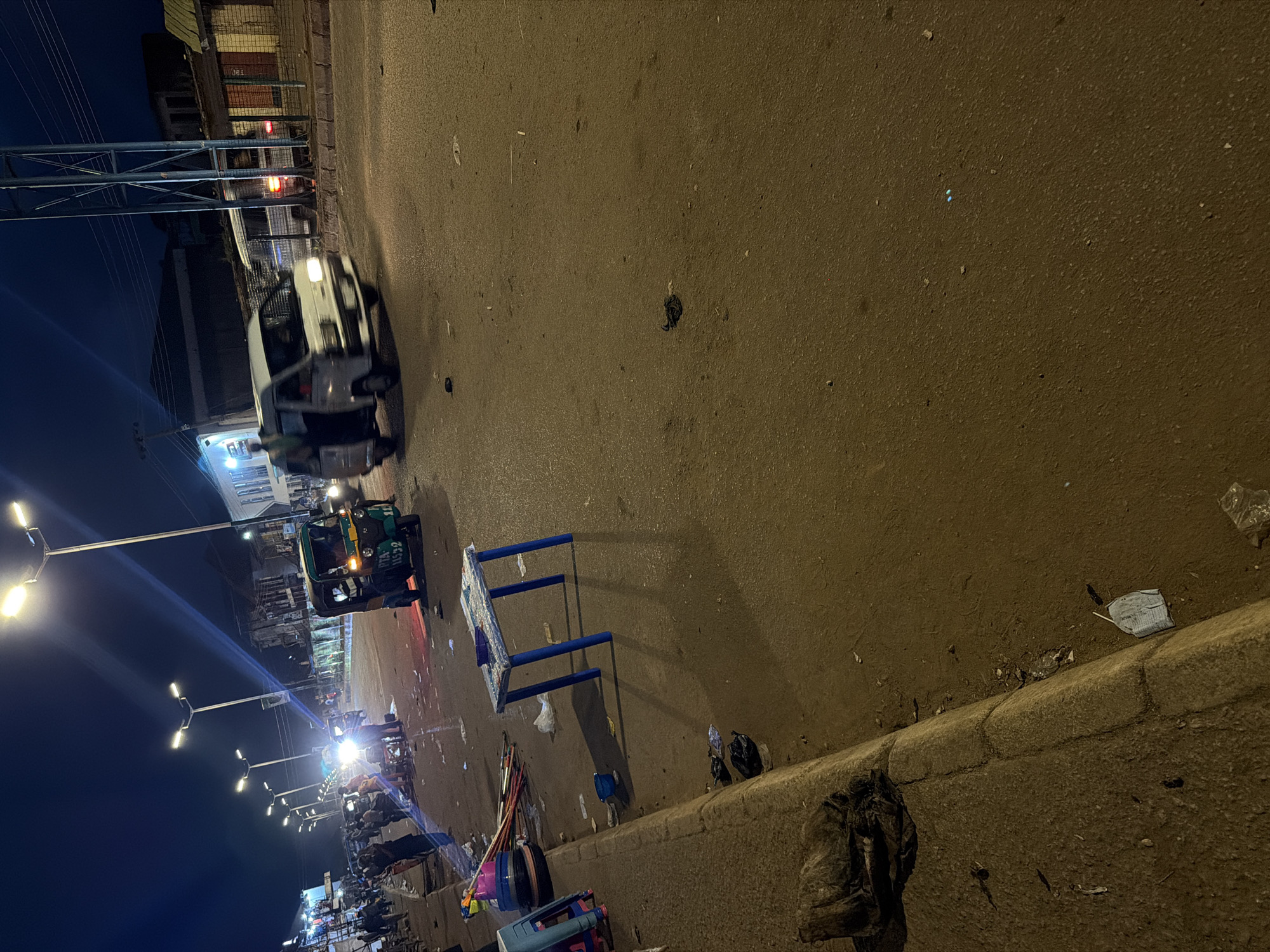 A nighttime scene captures a street in Bukuru, Nigeria, illuminated by streetlights and vehicle headlights. The road is wet, reflecting the artificial lights, and strewn with litter, including plastic bags, debris, and a fallen blue plastic table with white tape on its surface. In the foreground, a curb runs along the left side of the street, with trash bags and other refuse piled near it. A stack of colorful plastic bowls and some long, thin objects, possibly sticks or poles, are also visible on the side.
Further down the street, a three-wheeled auto-rickshaw, painted green and yellow, is stopped or moving slowly. A white van is also on the road, its headlights on, and a person can be seen in its open side door. Other vehicles, including what appears to be another van, are present in the background. Several people are standing on the sidewalk and near the vehicles, suggesting activity or a gathering. Buildings line the sides of the street, some appearing commercial with illuminated signs. The overall atmosphere is one of a busy urban environment at night, with evidence of recent activity and a significant amount of litter on the ground.