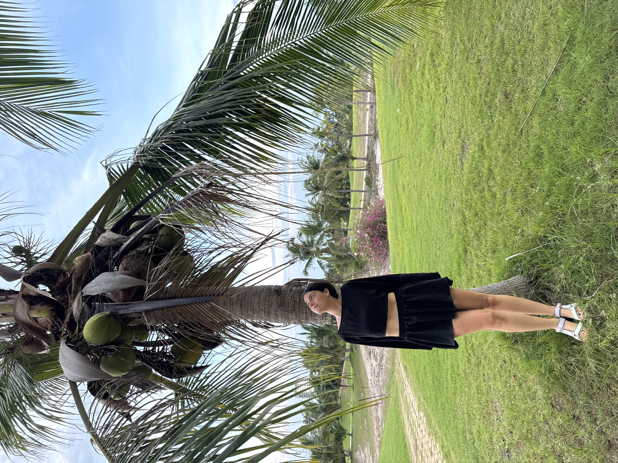 A young woman is standing next to a coconut tree in Phú Thủy, Vietnam. The woman is wearing a black, two-piece outfit with white sandals. The background shows a grassy area and other palm trees. The sky is blue with some clouds. The tree has several green coconuts. The photo has been rotated 90 degrees counterclockwise.