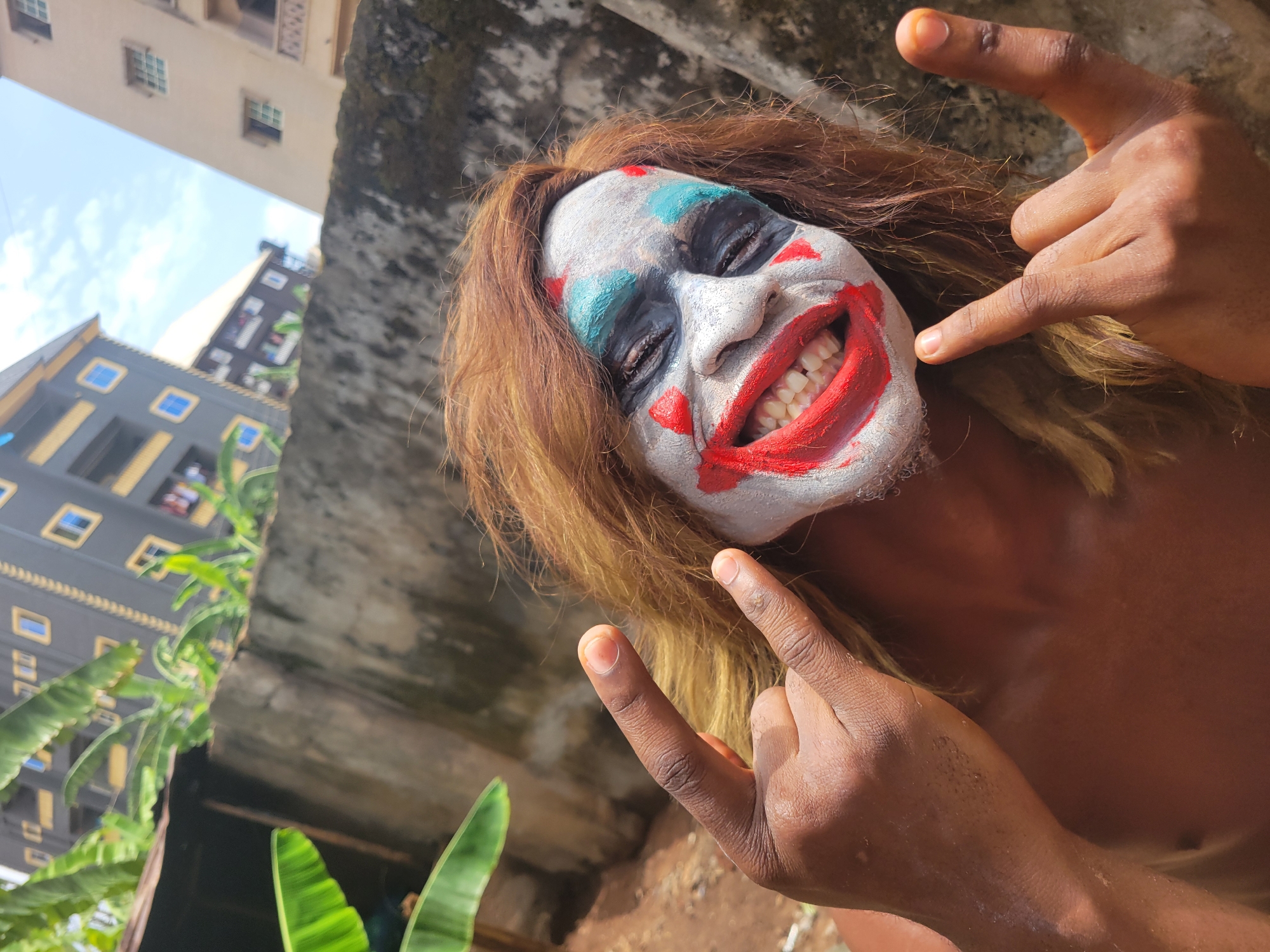 A person is shown from the chest up, angled upwards, with the tops of buildings and trees visible in the background. The person has brown hair and is wearing clown makeup with white face paint, red lips, and blue and red marks around the eyes. They are smiling widely, showing their teeth. The person is making the "rock on" hand gesture with both hands. The location appears to be outdoors, possibly near a city or town with a mix of concrete structures and greenery.