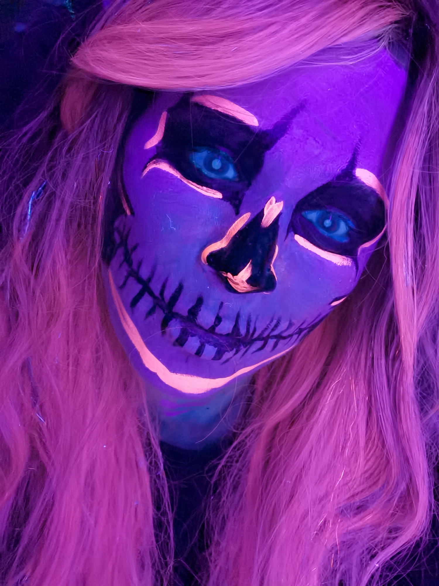 A close-up, low-angle photograph shows a person with pinkish blonde hair and skull-themed makeup, likely for a costume or special occasion. The makeup is predominantly purple with black accents around the eyes, nose, and teeth. Lighter pink highlighting outlines the skull shape. The person has light blue eyes. The overall lighting is vibrant and intense, suggesting the use of UV or neon lighting to enhance the makeup's glow. No discernible location or additional contextual clues related to the city of Chaikovsky, Russia, are present in the image.