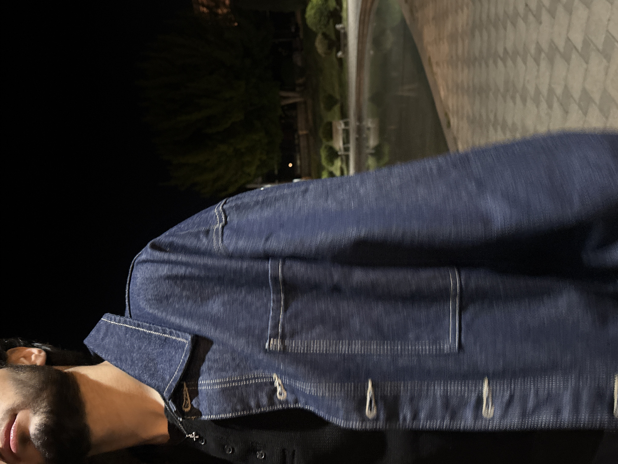 The image shows a person wearing a blue denim jacket at night. The background features a paved area, trees, and a body of water, possibly a canal or decorative pond. Based on the style of the jacket and the architectural elements, the location could be in Yerevan, Armenia, as requested.