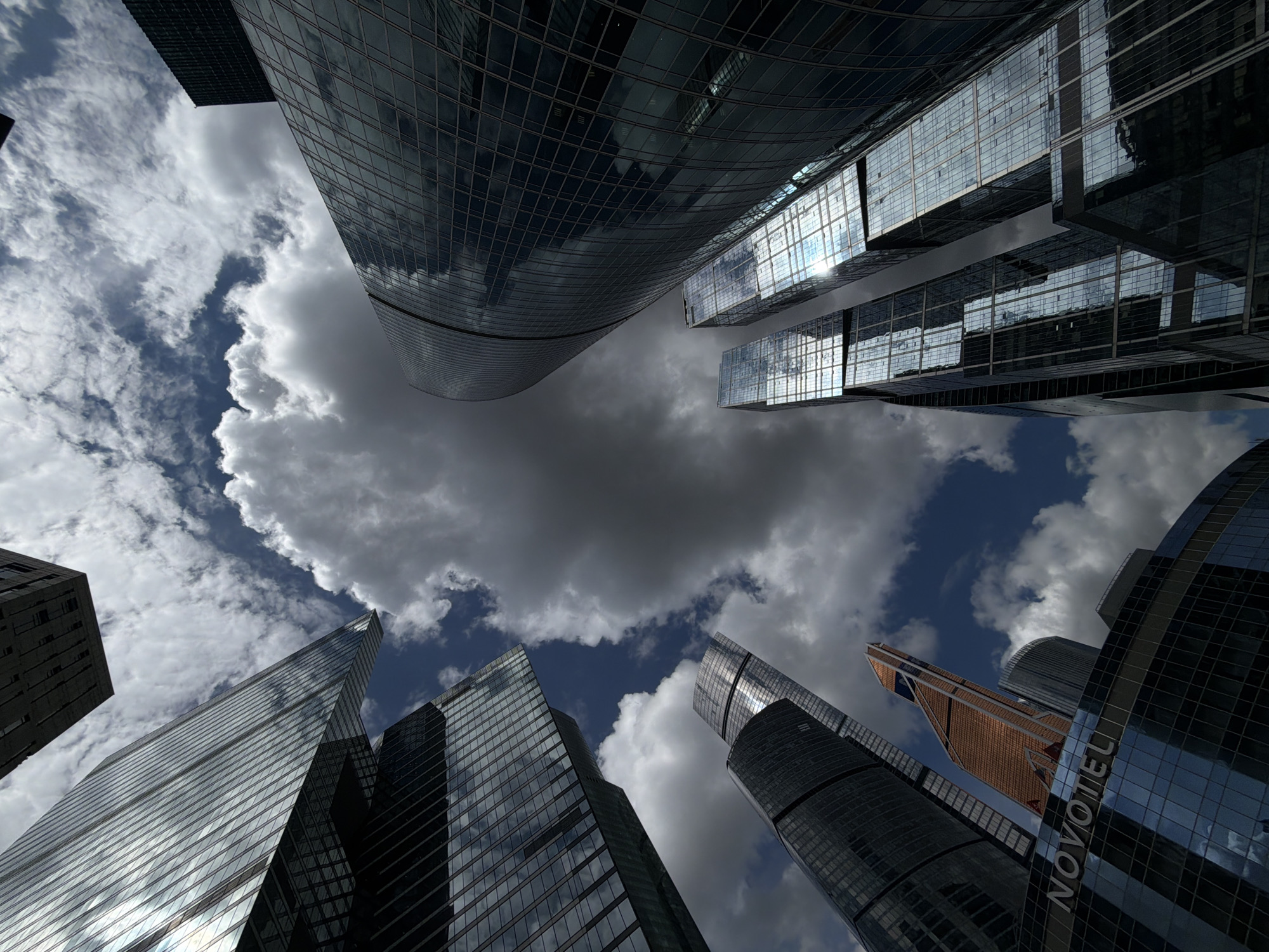 The image is a low-angle shot of several skyscrapers in Khimki, Russia, viewed from the ground looking upwards. The buildings are primarily made of glass and steel, reflecting the sky and clouds. A large, grey cloud formation dominates the center of the image, partially obscuring the sky. The sky visible around the cloud is blue with smaller, white clouds. The composition of the image emphasizes the height and scale of the buildings and the dynamic interplay between architecture and the natural environment. The word "NOVOTEL" is visible on one of the buildings.