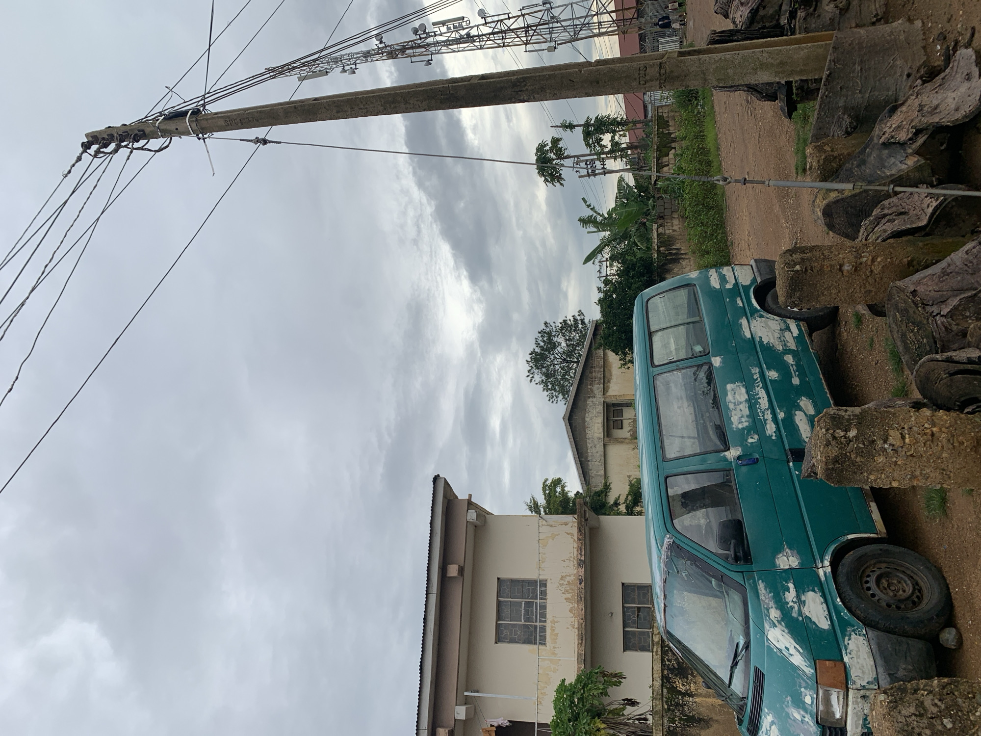 A teal-colored Volkswagen Transporter van, with significant paint peeling and white primer showing through, is parked on a dirt road in Rafin Par, Nigeria. To the left of the van is a two-story, light-colored building with a metal roof and barred windows. Behind the van and to the right, a tall concrete utility pole stands, supporting numerous overhead wires. Further to the right, a large metal telecommunications tower with multiple antennas is visible. The environment is overcast with heavy clouds in the sky. In the background, several trees and other buildings can be seen. Two individuals are walking in the distance near the telecommunications tower. In the foreground, several large, rough-hewn logs and stone posts are present.