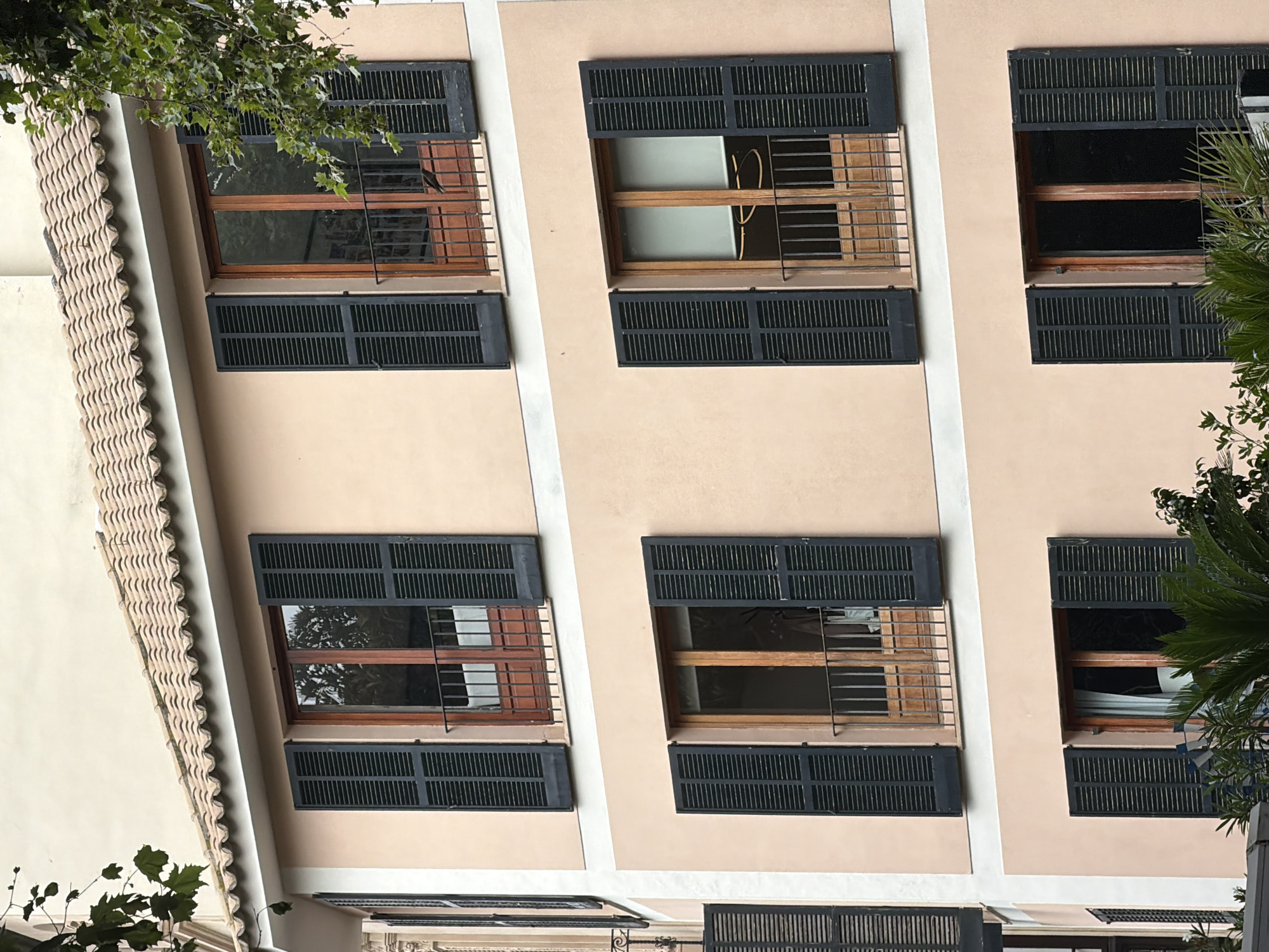 The image shows the facade of a building in Palma, Spain. The building has multiple stories with peach-colored walls and dark gray shutters on the windows. The windows themselves are framed in wood. The topmost part of the building is topped with a terracotta tile roof. There are some trees and foliage visible on the right side of the image, obscuring part of the building. The windows appear to have small balconies with metal railings. One of the windows on the second floor from the top has a bird perched on its railing. The image is taken from a low angle, looking up at the building.