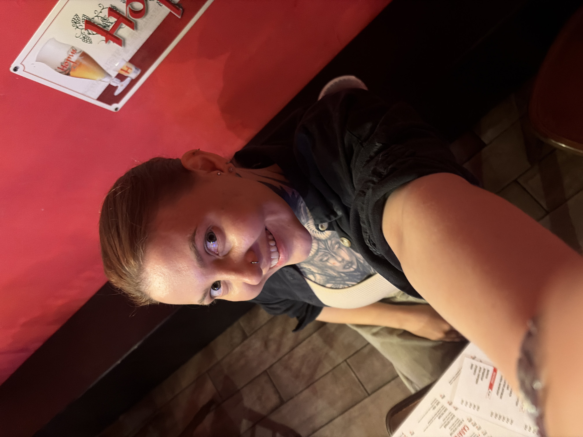 A high-angle shot shows a person sitting at a table. The person is looking at the camera with a wide smile and has a nose ring. They are wearing a black shirt over a light-colored top and have tattoos on their chest and arm. The background is dark, with a red wall visible on the left side of the frame. A sign with an image of a beer glass and the word "Hofa" is hanging on the red wall. The table has a menu on it. The scene appears to be indoors, possibly a restaurant or bar. The lighting is dim, suggesting it could be evening. The floor is tiled with a brick pattern.
