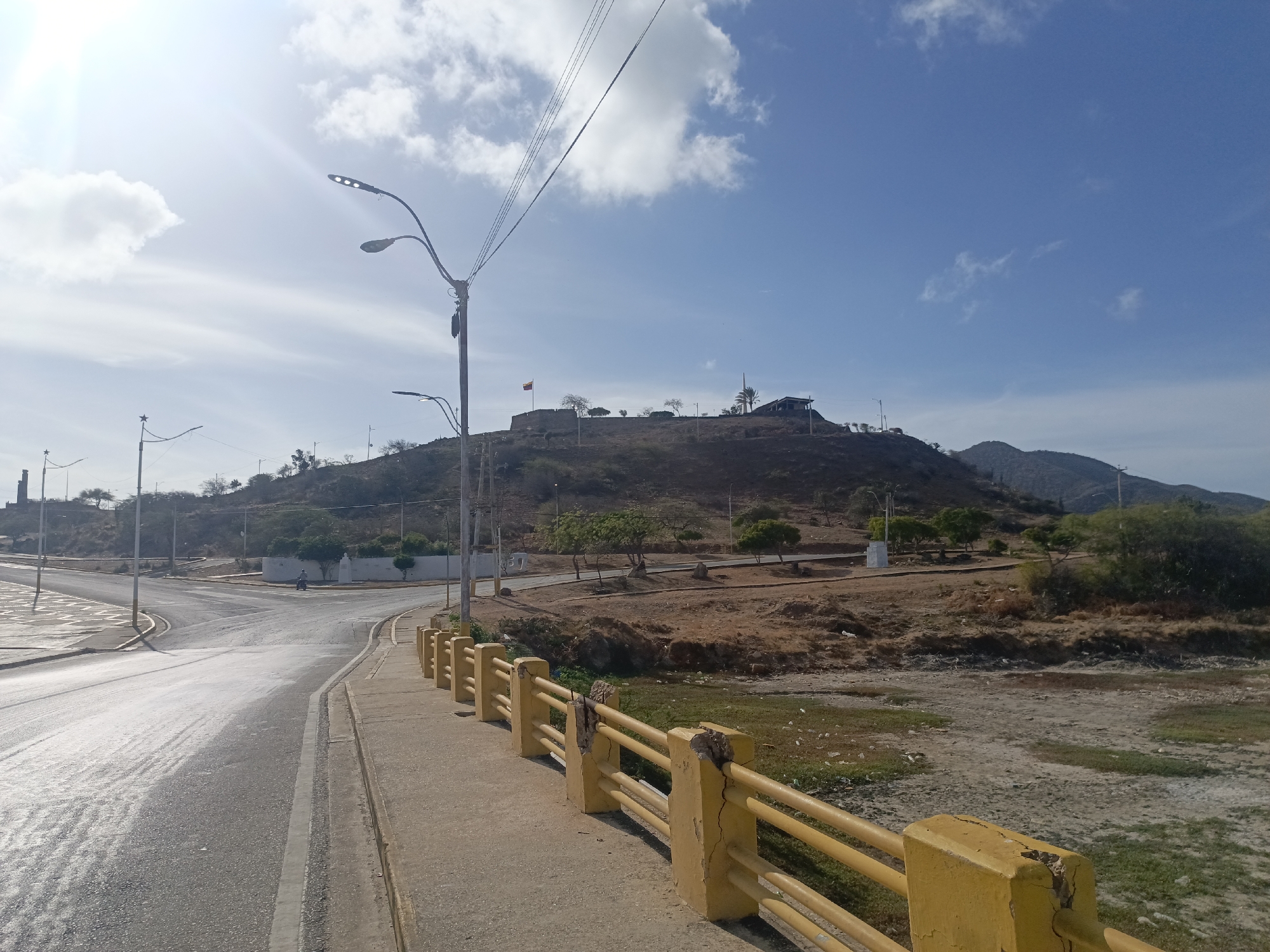 The image depicts a daytime scene in Juan Griego, Venezuela. A paved road with a sidewalk and a yellow railing is in the foreground. A dry riverbed with some green vegetation is visible beyond the railing. In the background, a hill rises with a structure and a flagpole flying a flag on its summit, suggesting a historical site or monument. Utility poles and power lines are visible in the sky. The sky is mostly blue with some scattered white clouds. There are some trees and sparse vegetation on the hill, and additional hills are visible in the far distance. The overall impression is of a sunny day in a coastal or semi-arid environment.