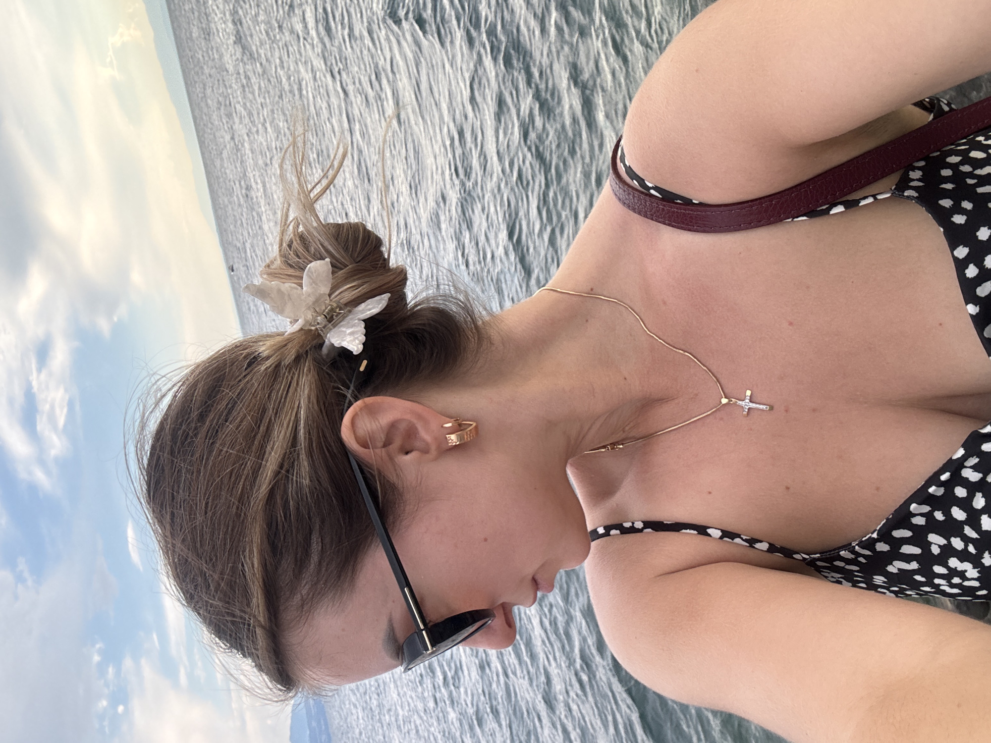 A young woman is captured in a profile shot, looking down and to the left. She is outdoors, with a body of water and a cloudy sky in the background, suggesting a lakeside or coastal location. The woman has her hair pulled up in a bun and is accessorized with sunglasses, gold hoop earrings, and a gold necklace with a cross pendant. She is wearing a black and white polka-dot dress. A dark red purse strap is visible over her shoulder. The overall scene evokes a sense of casual leisure by the water.