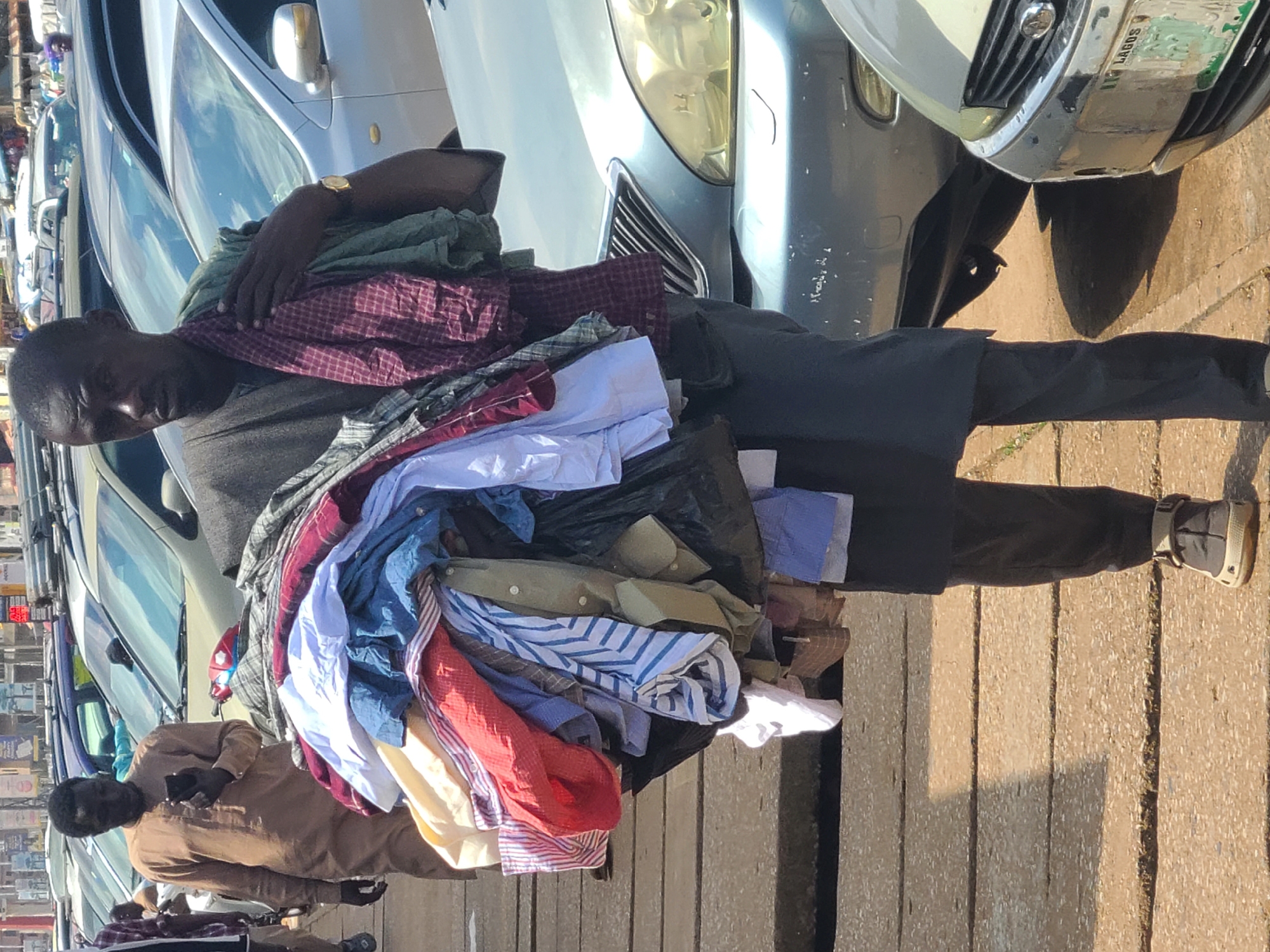 The image shows a man in Jos, Nigeria, carrying a large bundle of clothes on his back, draped over his shoulder and chest. He is wearing a dark grey t-shirt and black trousers. His head is bowed, and his face is partially obscured by the clothes. A gold watch is visible on his left wrist.
The man is standing on a paved sidewalk next to a row of parked cars. The cars are of various colors, including silver, light blue, and a darker blue. One of the cars has a license plate that reads "LAGOS". The scene appears to be outdoors, possibly in a marketplace or a street with vendors. The lighting suggests it is daytime, with sunlight casting shadows on the ground.
The bundle of clothes includes various types of shirts and fabrics in different colors and patterns, such as red, white, blue, and green. Some of the shirts are folded, while others are loose. The way the man carries the load suggests he is a vendor or transporting goods.
Another person, dressed in a light brown traditional outfit, is visible to the left of the main subject, holding a smartphone. In the background, more cars and some blurry figures can be seen, indicating a busy urban environment. The overall impression is of a bustling street scene in a Nigerian city.