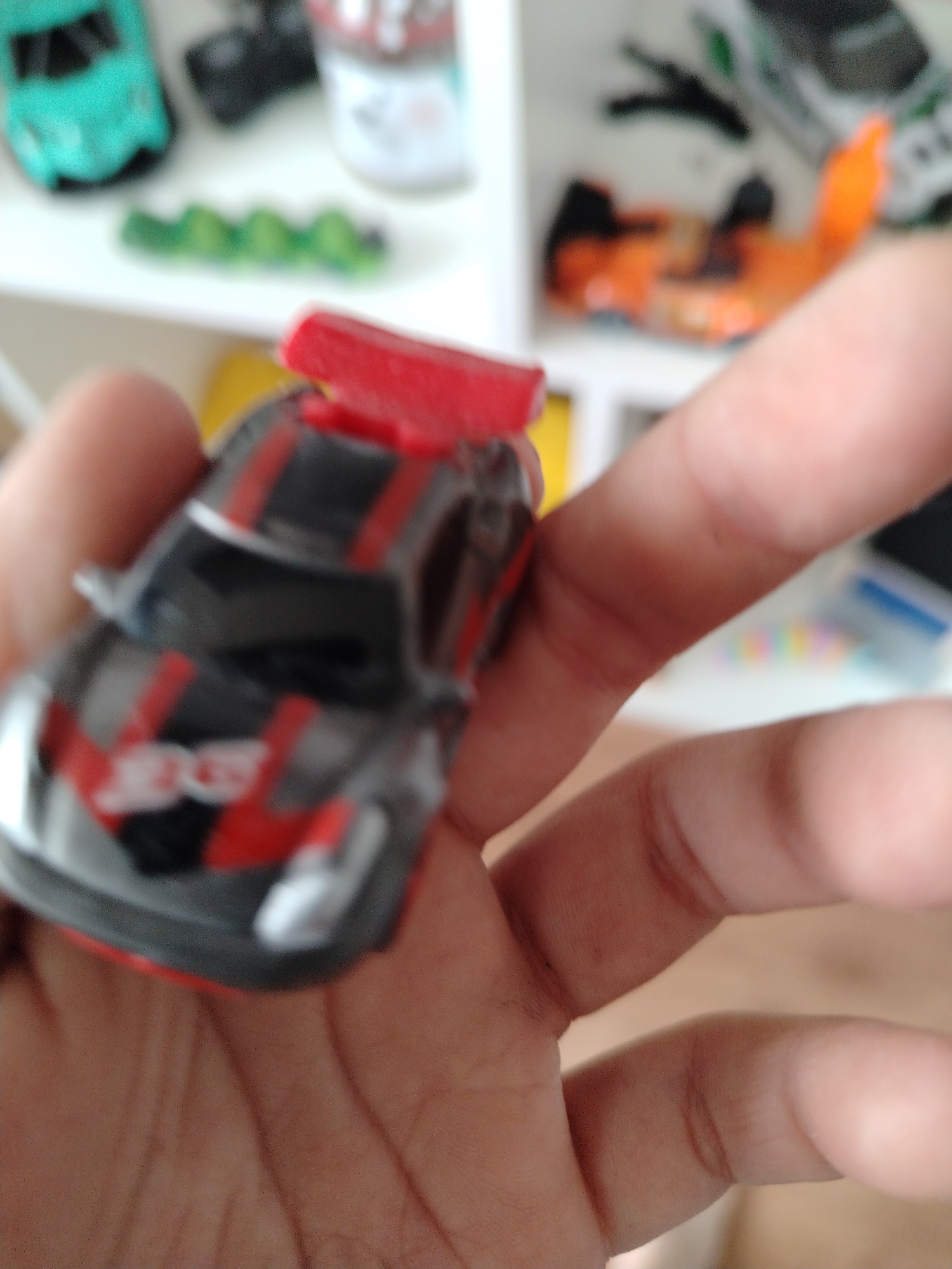 An out-of-focus, close-up shot captures a hand holding a toy race car. The car is predominantly black with red accents and stripes. In the background, out-of-focus objects suggest a shelf or display, including what appears to be other toys and possibly a container with colorful beads. The lighting is bright, casting shadows and highlighting the textures of the hand and the toy. The setting is indoors, with a neutral background indicating a domestic or retail environment. The car has a spoiler and appears to be a miniature replica of a sports car.