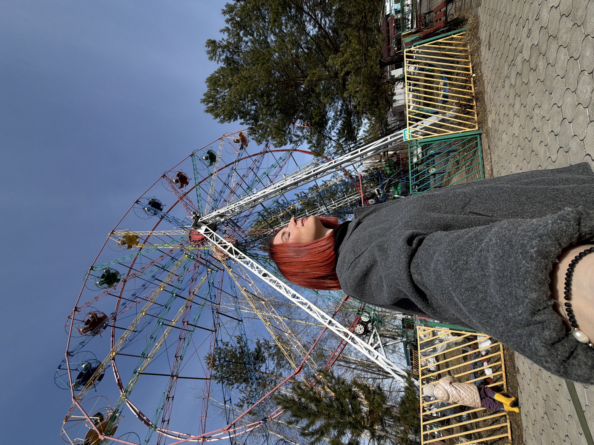 A medium shot features a person standing in front of a colorful Ferris wheel in Chaikovsky, Russia. The person is oriented vertically in the frame, with their head tilted back, gazing upwards. They have auburn hair and are wearing a gray jacket. The Ferris wheel, a prominent structure, is multi-colored, consisting of red, blue, yellow, and green carriages. The background shows a bright blue sky, some greenery from trees, and sections of a park fence that has yellow and green bars. A paved walkway is visible in the foreground.