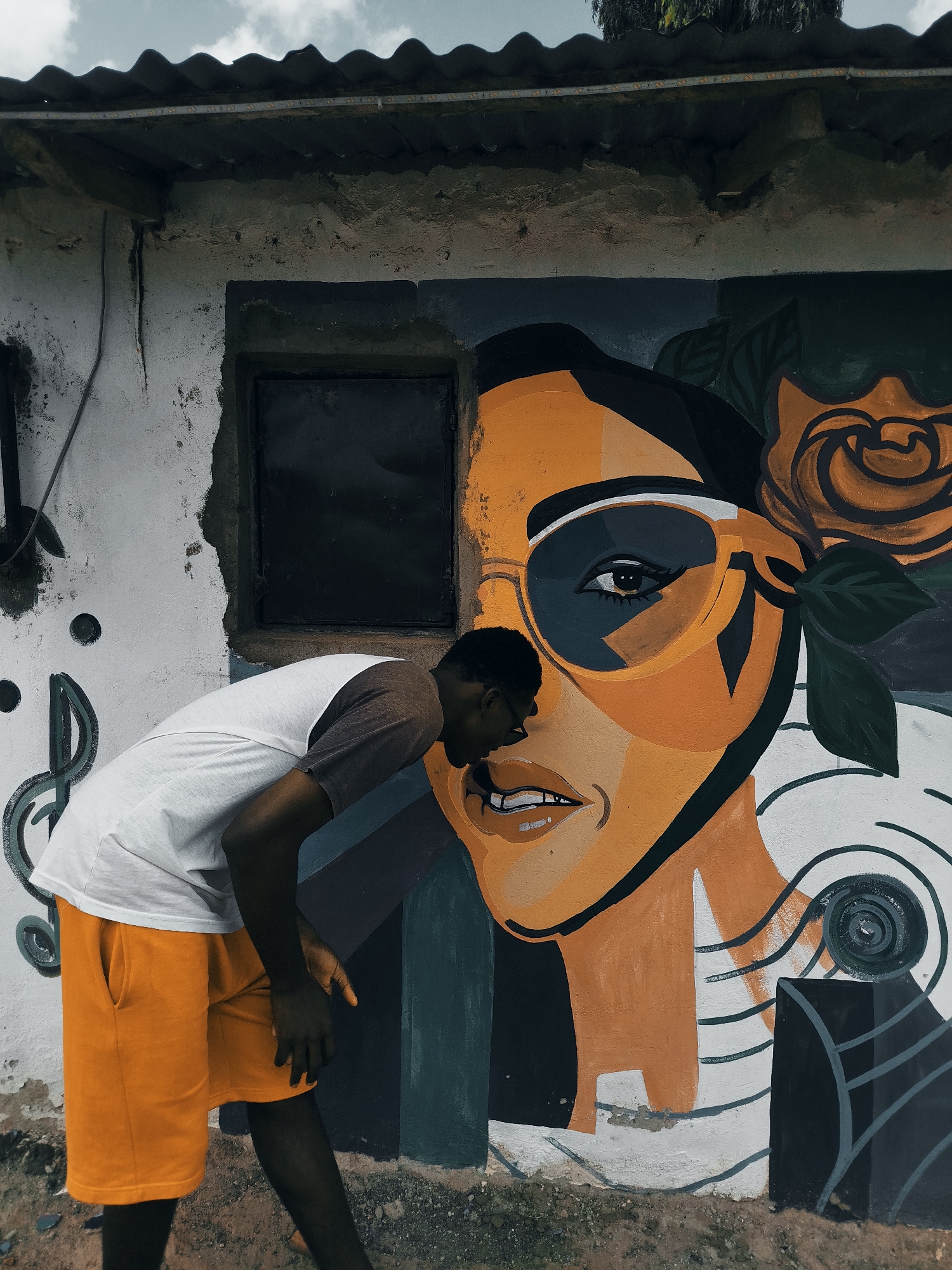 A young Black male with dark skin and short black hair, wearing a white t-shirt with dark gray sleeves and bright orange shorts, leans in towards a large mural painted on a weathered white concrete wall. The mural depicts a stylized portrait of a woman with orange skin and large, dark-rimmed sunglasses with silver reflections. A large orange rose with dark green leaves is positioned behind her head. Musical notes are painted on the wall to the left of the mural. The building's roof is corrugated metal, and the sky is overcast. The scene appears to be an outdoor setting in Narayi, Nigeria, indicated by the distinctive architecture and environment. The male is positioned with his head close to the mural, as if examining it. The wall is chipped and worn in places, suggesting age and exposure to the elements.