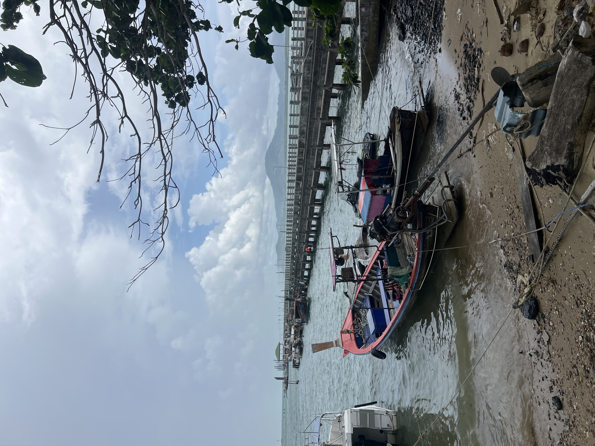 The image depicts a serene coastal scene in Pa Khlok, Thailand, with several longtail boats moored near a weathered concrete pier that extends into the calm, choppy sea under a cloudy sky. The foreground shows a sandy and rocky shoreline littered with driftwood, ropes, and debris. Two prominent longtail boats, one red and blue with an outboard motor, and another smaller one with a pink and blue canopy, are tied to the shore. The pier, with its sturdy concrete pilings and railings, stretches across the water, leading to a distant structure with a green roof. In the background, lush green hills rise against the horizon under a sky filled with fluffy white clouds. The overall atmosphere is one of tranquil seaside life, typical of the region.