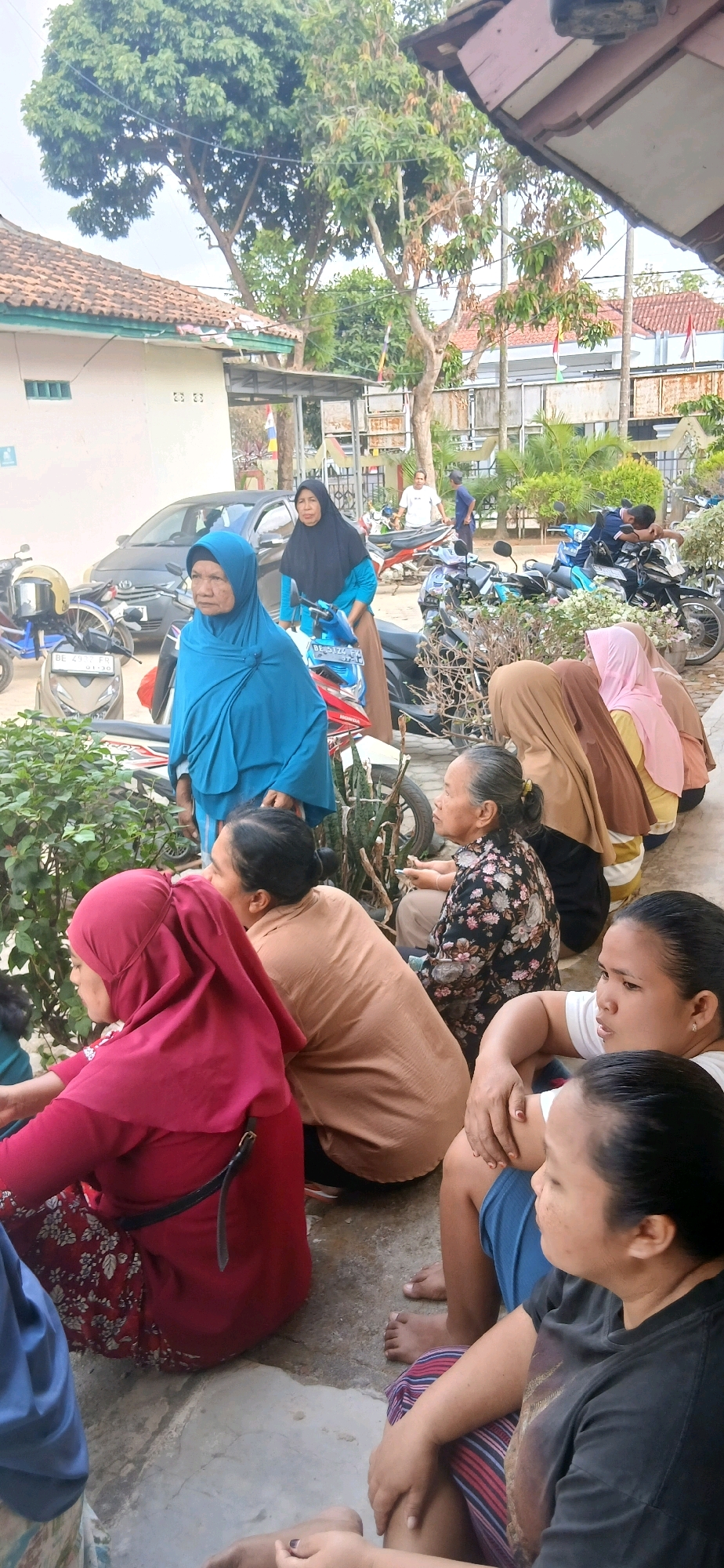 The image depicts an outdoor scene, possibly in a residential area of Metro, Indonesia, where a group of women are gathered. In the background, several motorcycles are parked, suggesting a public or communal space. A car is also visible parked behind a woman wearing a blue hijab. The women are dressed in various headscarves and modest clothing. Some are seated on the ground, while others are standing. The overall impression is of a casual gathering or waiting area. The environment appears to be somewhat rural or suburban, with vegetation and buildings visible. The lighting suggests it is daytime.