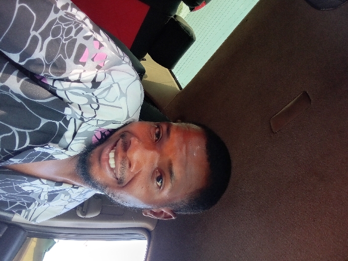 A smiling man with a beard, wearing a patterned gray and black shirt with pink accents, is angled to the left in the frame. His eyes are open and directed at the viewer, showing his teeth. The photo appears to be taken from inside a vehicle, with the interior ceiling behind him. A car window and part of a seat are visible in the background.