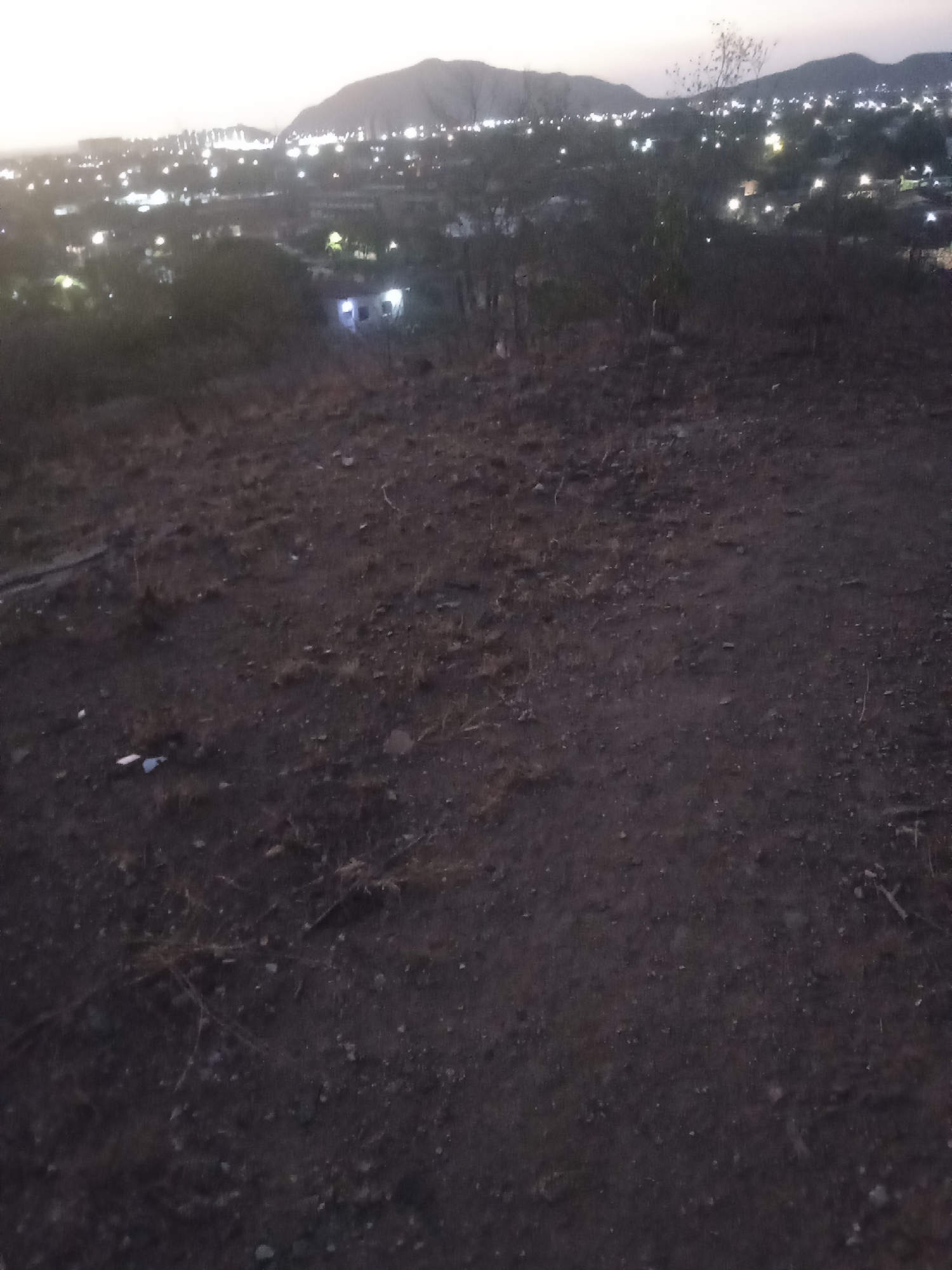 The image shows an elevated, barren foreground, possibly a hilltop or a slope, covered in dark soil with scattered rocks and sparse vegetation. In the distance, there is an urban area, potentially Juan Griego, Venezuela, with numerous illuminated buildings and streetlights visible against the dark sky. There are mountains silhouetted in the background.