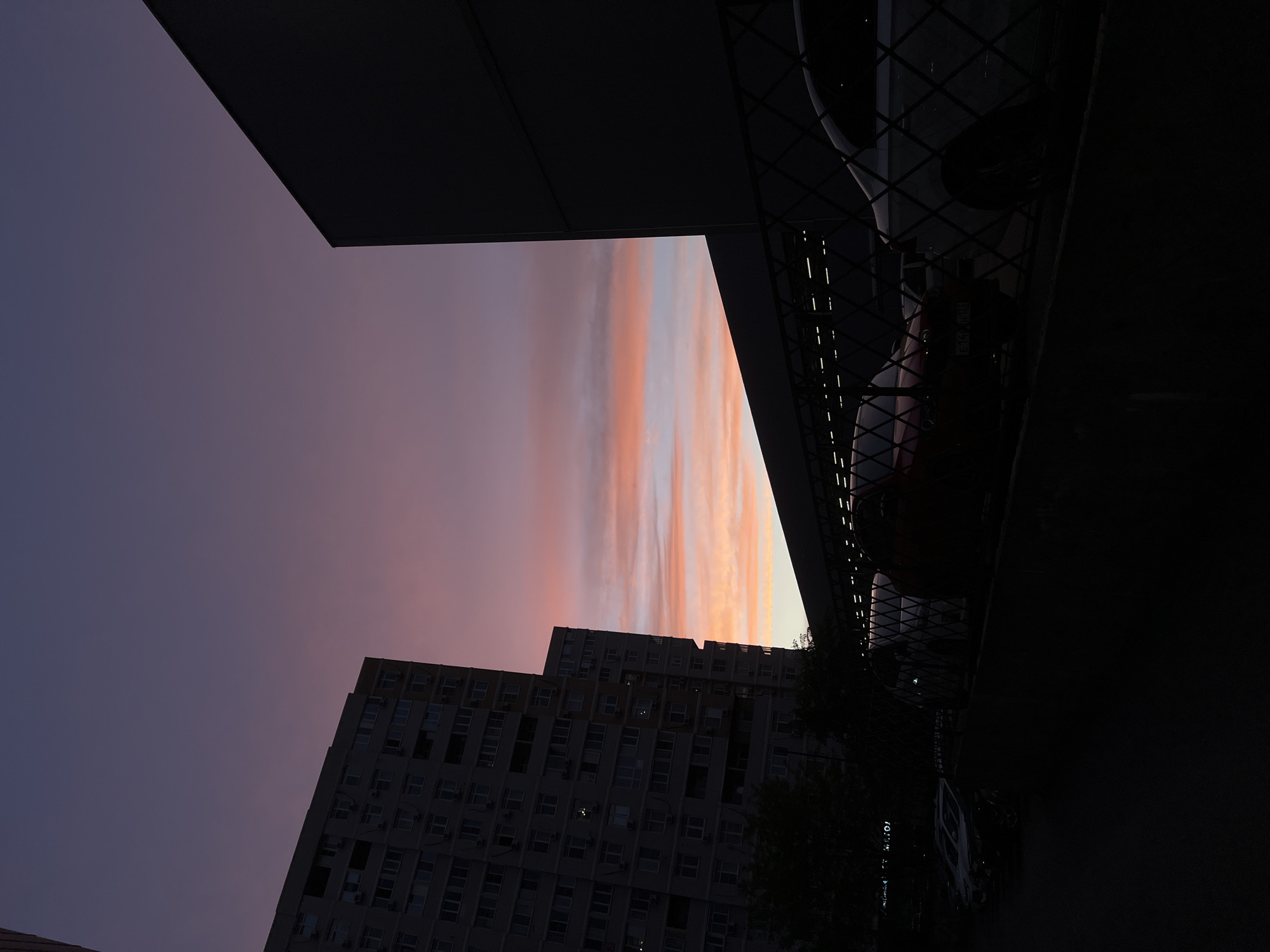 The image depicts a view of the sky at dusk, framed by buildings and structures in Krasnodar, Russia. The sky exhibits a gradient of colors, transitioning from purple-blue to orange-pink, indicative of a sunset or sunrise. Two high-rise buildings are visible in silhouette against the colorful sky, their windows indicating a densely populated structure. A dark, angular structure occupies the upper left corner of the frame, while a metal fence or grate with vehicles behind it is visible on the right. The lighting suggests low-light conditions, possibly after sunset.