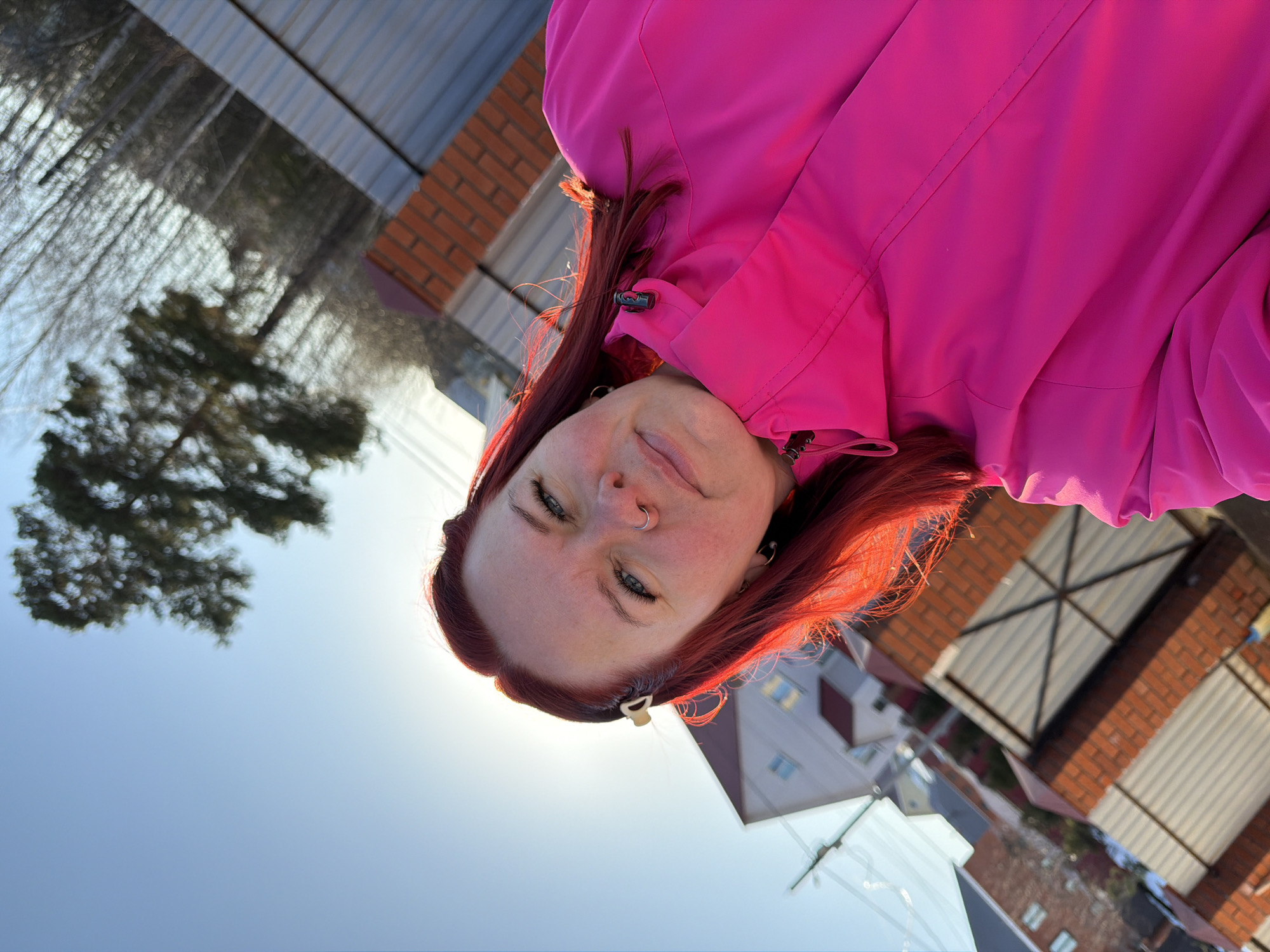 A medium shot, taken from a low angle, depicts a woman with fair skin and red hair wearing a bright pink jacket. She has a nose ring and is looking down at the camera with a slight smile. A small hair clip is visible in her hair near her temple. The background shows a bright blue sky, a tree with bare branches, a brick wall, and several buildings. This photo appears to be taken outdoors in a residential area in Chaikovsky, Russia.