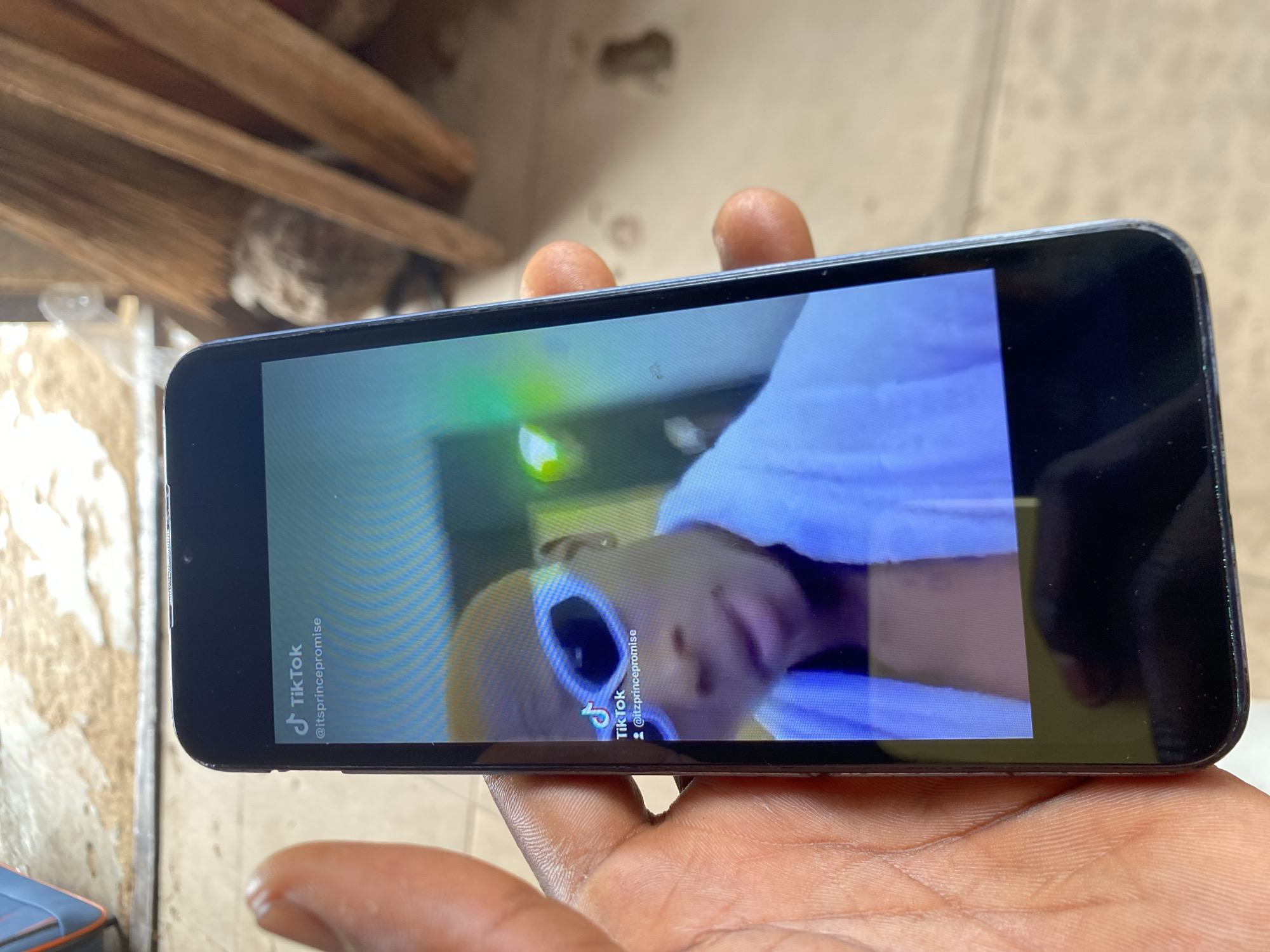 A hand holds a black smartphone, the screen displaying a TikTok video. The TikTok video features a person wearing large, white-framed sunglasses and a white garment. Text on the video reads "@itzprincepromise." The background suggests the image was taken outdoors, potentially in Bukuru, Nigeria, indicated by the urban or semi-urban environment visible. Other objects around the hand include wood planks in a building.