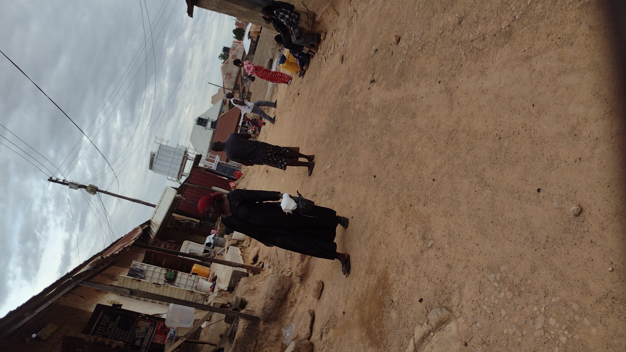 The image shows a dirt road in Jos, Nigeria. The scene includes people walking along the road and some sitting on the side. There are buildings on one side of the road, one appears to be a shop. Power lines are visible overhead. The sky is cloudy.