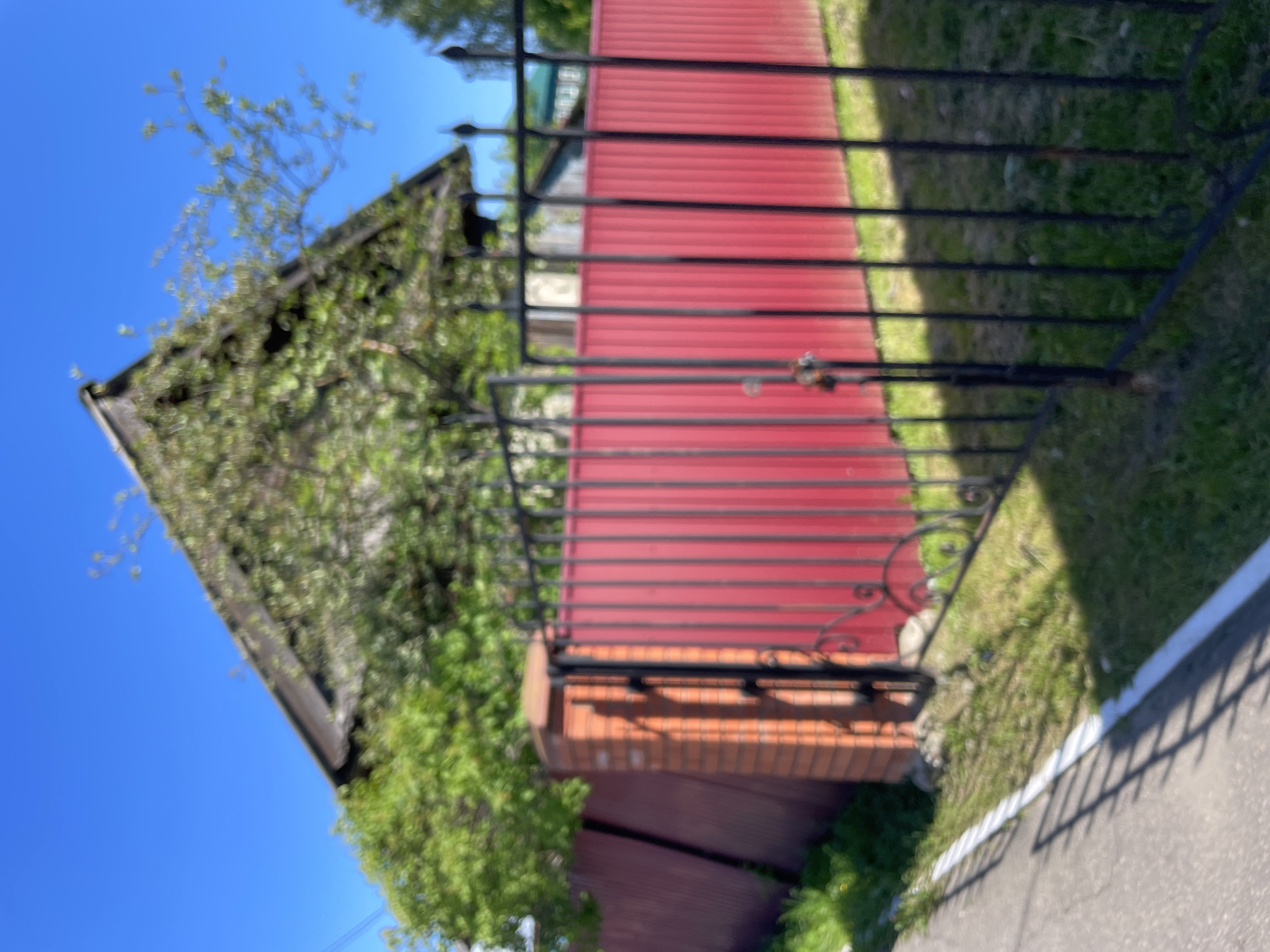 A daytime, outdoor, eye-level shot captures a residential scene in Krasnye Baki, Russia. In the foreground, a black wrought-iron gate stands partially open, revealing a bright red metal fence behind it. The fence extends to the left, where it meets a brick pillar. To the left of the gate and fence, a paved walkway with a white line runs along its edge. Behind the red fence, a house with a dark, sloping roof and partially covered in green foliage is visible. Lush green trees and grass surround the property. The sky above is a clear, bright blue.