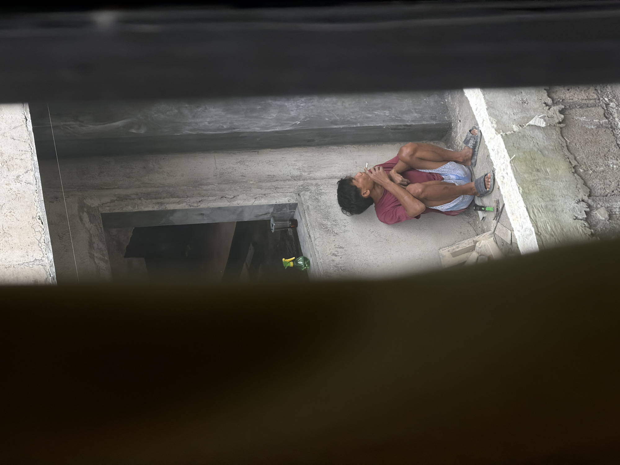 An overhead shot, taken through a narrow opening or from behind an obstruction, shows a man taking a break at a construction site. The image is rotated 90 degrees clockwise, presenting the man lying on his side. He is seated on a concrete floor, with his knees drawn up, wearing a maroon short-sleeved t-shirt, blue and white patterned shorts, and black slide sandals. He holds a lit cigarette to his mouth, with smoke or vapor visible. His expression is neutral as he looks upwards (relative to the image orientation).
The setting is an unfinished concrete structure, with rough grey walls and floor. In the foreground, near the man's feet, a hammer with a green handle and another hand tool, possibly a trowel or chisel, are visible on the concrete. A pair of black flip-flops or sandals also rests on the ground. Further back, on a raised concrete surface, a green spray bottle with a yellow nozzle can be seen. A thin white string or line, likely a construction guide, is visible hanging near a concrete column on the left side of the frame. The scene suggests a daytime setting within a somewhat shadowed area of the building. No visible text is present. The general context points to a construction worker resting in North Kuta, Indonesia.