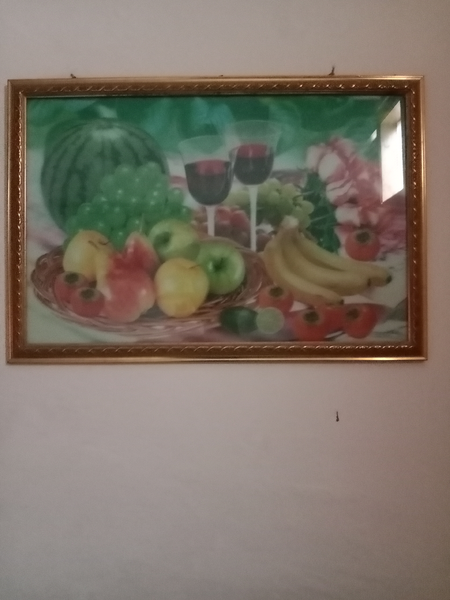 A framed still life painting of fruit and wine hangs on a plain, light-colored wall. The painting features a watermelon, grapes, apples, bananas, and what appear to be tomatoes and limes, arranged on a wicker platter. Two glasses of red wine are also depicted. The frame is gold-colored with an embossed pattern. A small, dark speck, possibly a spider or a piece of debris, is visible on the wall above the painting. The location is identified as Kutosari, Indonesia.