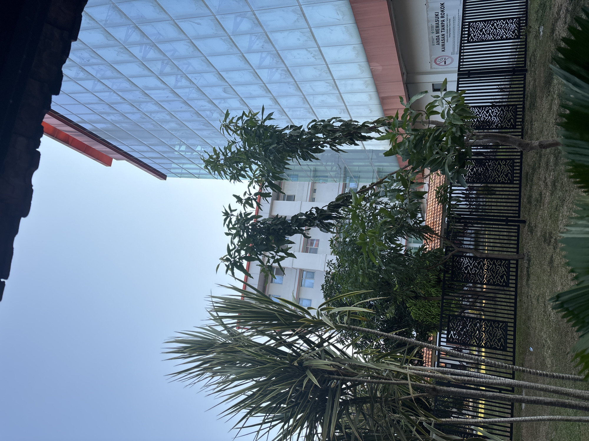 This image depicts the exterior of a modern building with a glass facade, possibly a hotel or office building, located in Cirebon, Indonesia. The building features a striking blue glass curtain wall and a red trim at the top. In front of the building, there is a black metal fence with decorative panels, separating the building from a grassy area. Several trees and large palm-like plants are visible in the foreground and middle ground, with lush green foliage. A sign on the building's exterior, visible through the fence, reads "ANDA MEMASUKI KAWASAN TANPA ROKOK" which translates to "YOU ARE ENTERING A NO SMOKING AREA," indicating it is a public or semi-public space. The sky is clear and blue, suggesting daylight.