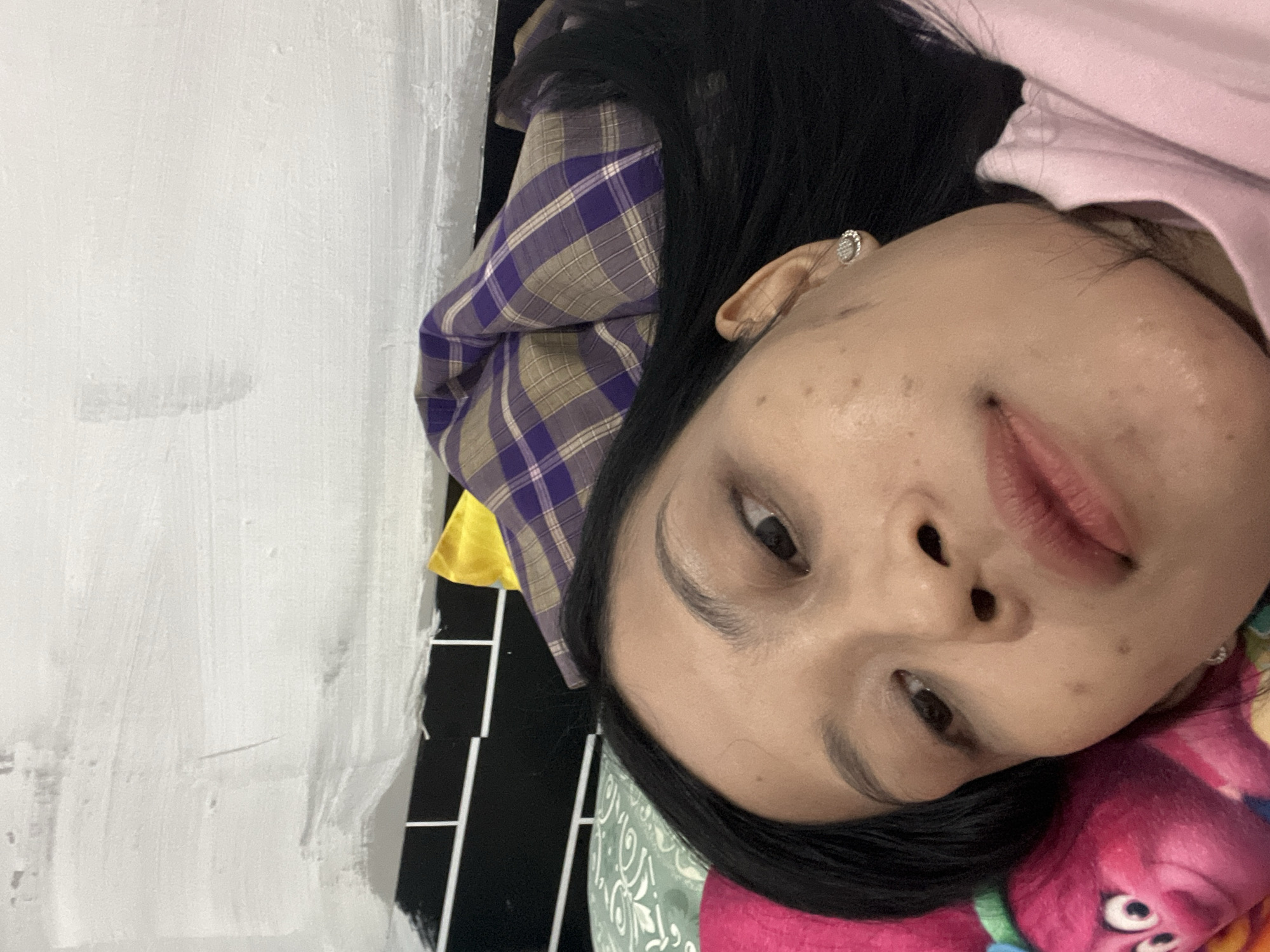 The image is a close-up selfie of a young woman lying down, with her head turned towards the camera. She has long, dark hair and is wearing a pink shirt and a stud earring. The background consists of a white wall with some textured paint strokes, a black tiled pattern, and various pillows. One pillow is green with a patterned design, another is pink and appears to be a stuffed toy character with large eyes and a smile. A purple and tan plaid fabric is visible behind her head. The scene is indoors, likely in a bedroom. The woman's facial features include dark eyes, defined eyebrows, and full lips. There are some small brown spots on her skin. The overall impression is that of a casual, personal moment captured in a domestic setting. There is no discernible information in the image that would indicate a specific location in the city of Yogyakarta, Indonesia.