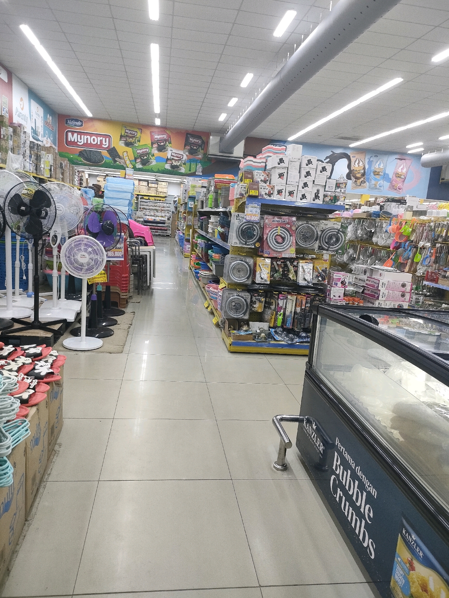 The image depicts the interior of a well-lit supermarket in Batam, Indonesia. The aisle features a variety of merchandise displayed on shelves and in freestanding units. In the foreground, to the left, are stacks of cardboard boxes with blue and white printing, topped with red and white flip-flops. Next to these, to the right, are several standing fans, including black, white, and purple models. Further into the aisle, red and pink plastic chairs are stacked. Shelves are laden with packaged goods, including what appear to be kitchenware, cleaning supplies, and food items. Some shelves are dedicated to items like gas hoses and lighters. To the far right, a large glass-fronted freezer or display case is visible, with signage that includes "KANZLER" and "Pertama dengan Bubble Crumbs." The flooring is tiled in a neutral color. The overall environment suggests a retail setting focused on household goods and groceries.