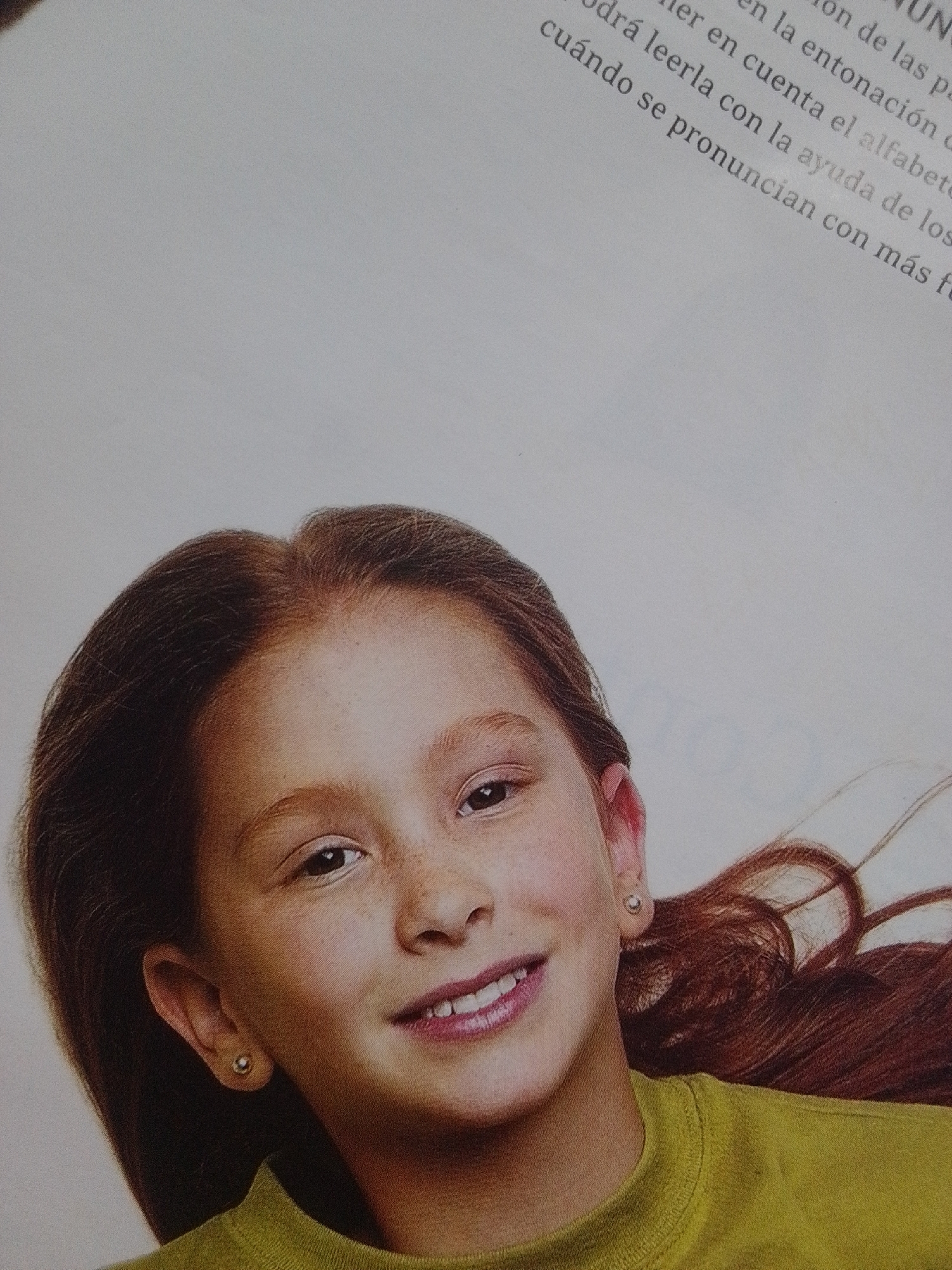 The image shows a close-up of a young girl's face and shoulders. She has brown hair, freckles on her cheeks, and is smiling with her mouth slightly open, revealing her teeth. She is wearing a green shirt and a small stud earring in her left ear. The background is a plain, light-colored surface, possibly a page from a book or magazine, with some text visible in the upper right corner. The text is in Spanish and appears to be related to pronunciation. There is no visible indication of the city of La Tagua, Venezuela, in the image.